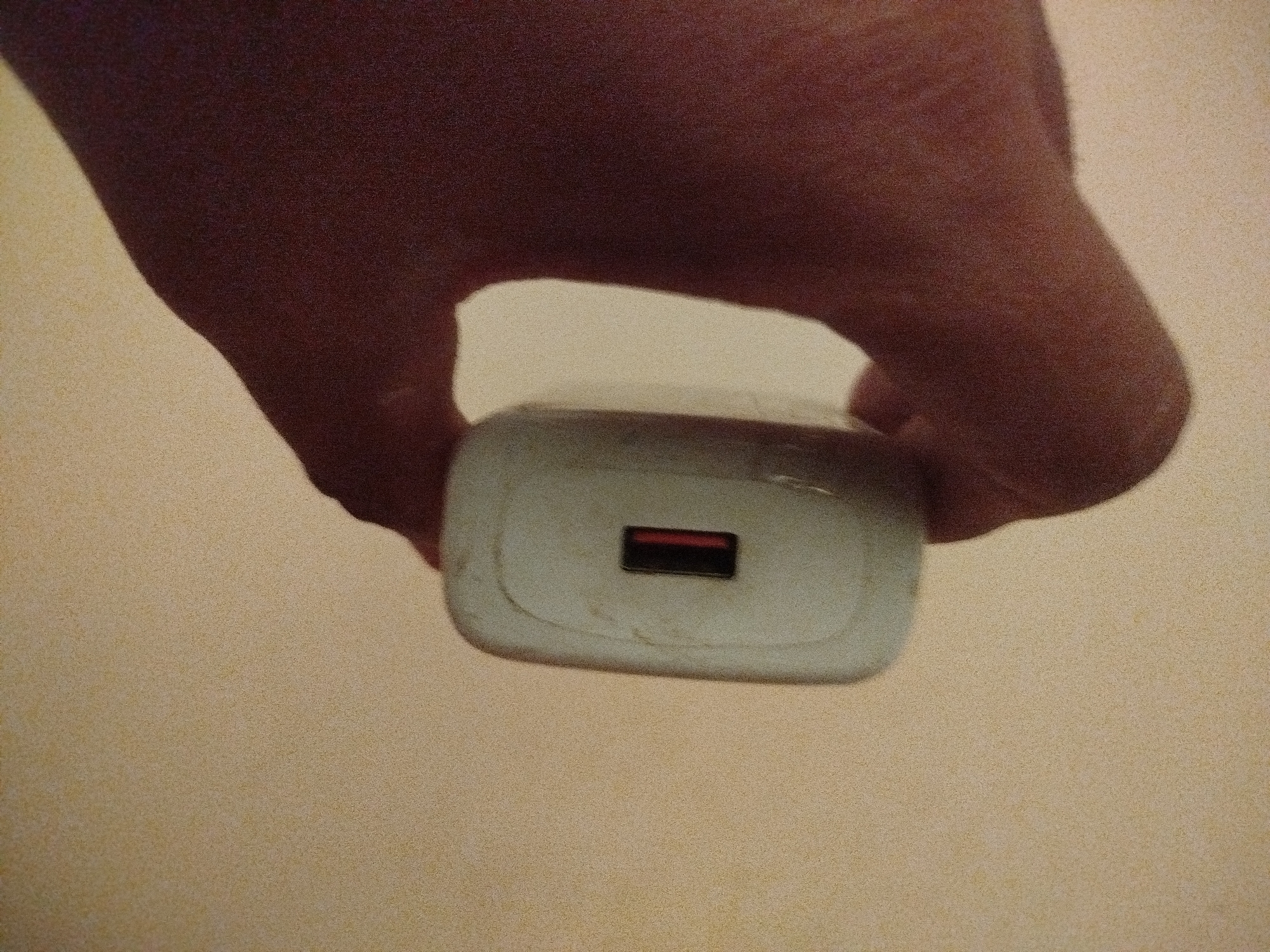 A hand is holding a white USB charger in the center of the image. The charger has a single USB-A port with a red rectangle visible inside. The hand is casting a shadow on a tan, textured surface. There is no indication of the city of Lassan, Pakistan in the image.