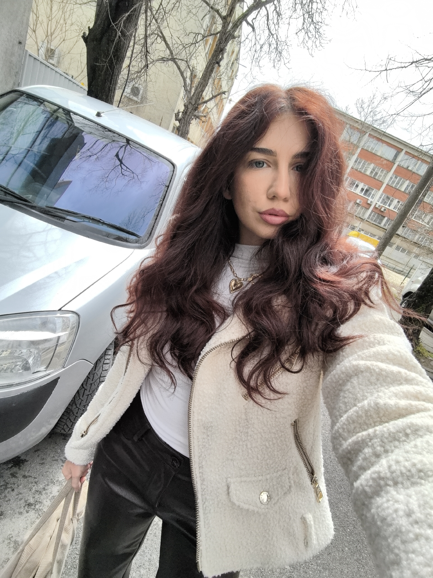 The image portrays a young adult female captured in a selfie on an asphalt surface in the city of Stara Zagora, Bulgaria. The subject has long, wavy dark auburn hair, blue-green eyes, and is wearing a white high-neck top, a white textured zip-up jacket with gold zippers and a gold button on the left pocket, and black leather-look pants. She wears a gold heart-shaped locket on a chain around her neck and holds a striped beige fabric bag in her left hand.
To the subject's left, a silver-grey van is parked, displaying its front hood, windshield, driver's side headlight, and a visible winter tire on the front wheel. The windshield reflects bare tree branches and the sky.
The background consists of urban buildings and bare trees. Behind the van, a light beige building with windows and an outdoor air conditioning unit is visible. To the right, behind the subject, a multi-story reddish-brown brick building with numerous windows and multiple external air conditioning units stands. Deciduous trees, devoid of leaves, are scattered throughout the scene, suggesting a late autumn or winter period. The sky is bright with partial cloud cover.