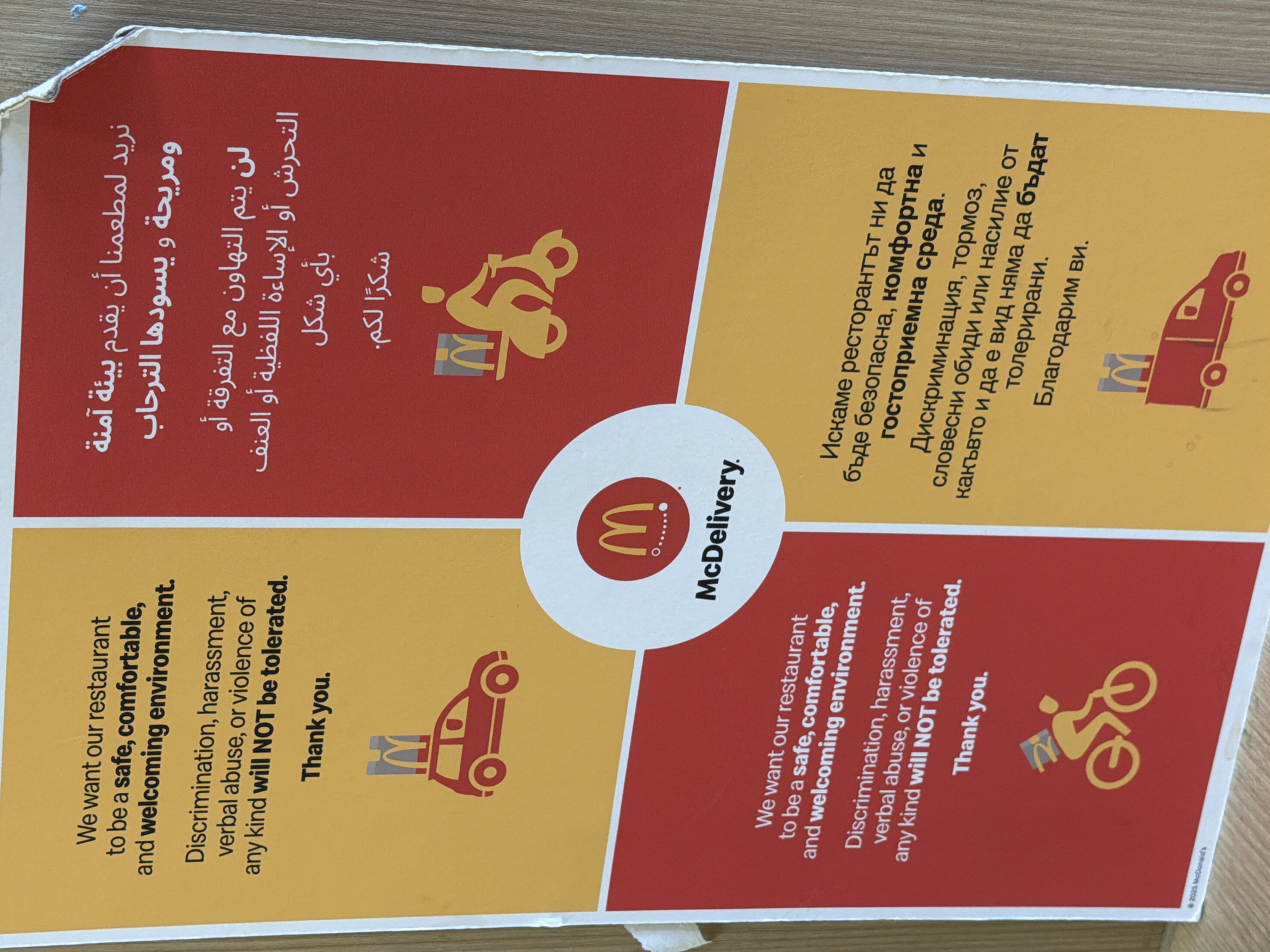 The image is a sign from McDonald's with a message about creating a safe and welcoming environment. The message is translated into three languages: English, Arabic, and Bulgarian. The sign states that discrimination, harassment, verbal abuse, or violence of any kind will not be tolerated. Each section has a McDonald's logo or icon and is separated by a circle in the center with the words McDelivery. The sign is located somewhere inside the restaurant or drive-thru.