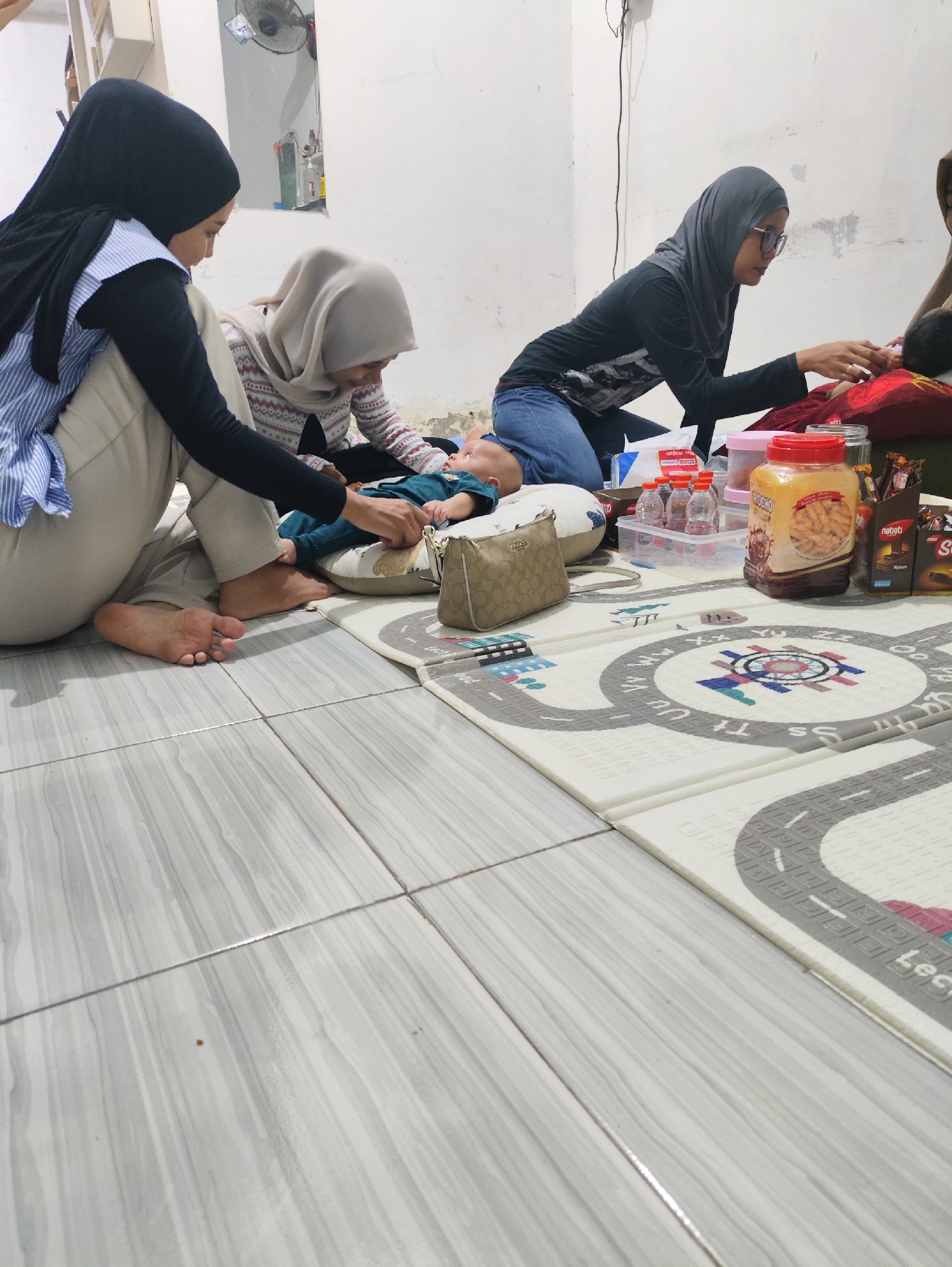 The image depicts an indoor scene with three individuals, two adult women and a baby, gathered on a mat on a tiled floor. The lighting appears to be artificial, creating soft shadows.
On the left, an adult woman wearing a black hijab and a blue and white striped shirt is seated on the floor, facing a baby. She is wearing light-colored pants. Her left leg is bent, and her right foot is visible.
In the center, another adult woman, also wearing a hijab (a light beige one), is kneeling. She is looking down towards the baby, who is lying on a white and gray mat with a road design. The baby is dressed in a teal-colored outfit.
To the right, a third adult woman wearing a dark gray hijab and glasses is kneeling. She is wearing a black long-sleeved shirt and blue jeans. Her hands are engaged in some activity with the baby or the items nearby.
Various objects are scattered on the mat and the floor around the individuals. These include a small beige handbag with a patterned design near the baby, several small bottles in a clear plastic container, a large jar of crackers with an orange label, a box of chocolates, and other packaged snacks. A pink lid is visible on one of the containers.
The environment is a room with white walls and a tiled floor. A ceiling fan is partially visible in the upper left corner of the image. Wires are also visible on the wall. The overall impression is a casual gathering, possibly a family moment, in a residential setting. The specific location in the city of Prague, Indonesia, is not determinable from the visual evidence alone.