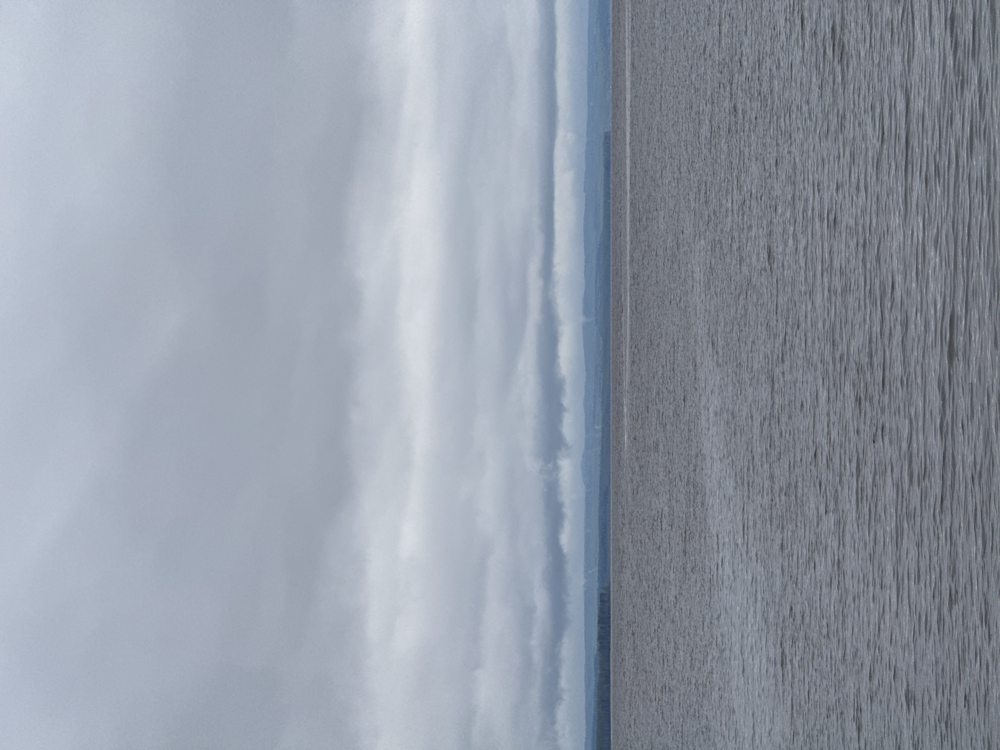 The image presents a landscape with an apparent division between sky and water. The sky occupies the left portion, displaying a range of grey hues indicative of cloud cover. To the right, the water surface is textured with vertical lines suggesting ripples or currents. The horizon line is indistinct, potentially obscured by mist or distance. The location is reportedly Chastyye, Russia, though there are no explicit landmarks or details to confirm this.