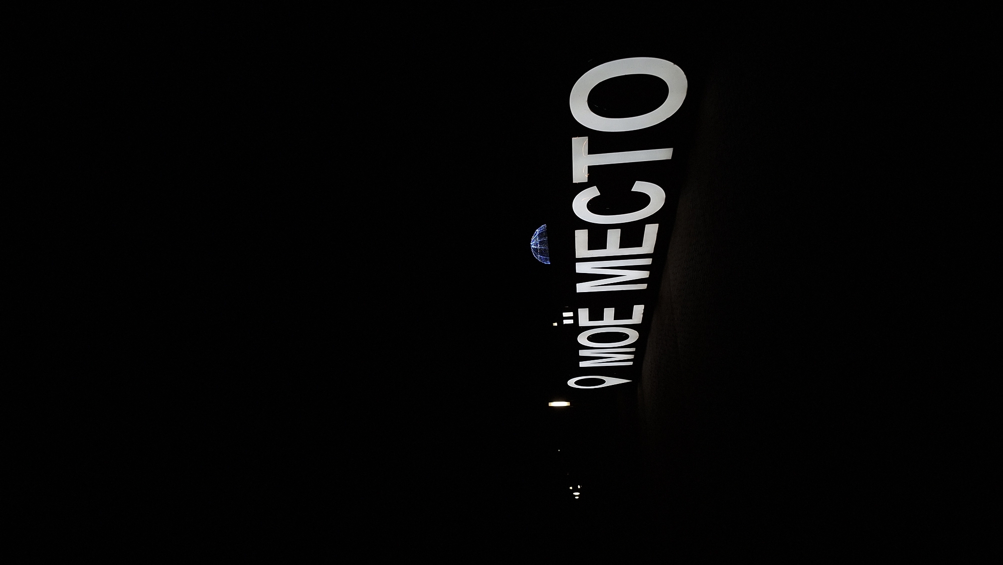 A low-angle shot captures a glowing, white sign at night, spelling out "МОЁ МЕСТО" (which translates to "My Place") in large, bold Cyrillic letters. The sign is positioned outdoors, with a dark, empty expanse behind it, suggesting a nighttime setting. To the left of the sign, a small, illuminated object resembling a stylized map pin is visible, further emphasizing the "place" theme. Above and slightly behind the text, a decorative, illuminated sphere or dome shape is suspended, casting a faint blue glow. The ground beneath the sign appears to be paved or tiled, with subtle lighting elements also visible in the distance to the left. The overall atmosphere is one of modern, ambient lighting in a public outdoor space, likely an urban area, though no specific architectural details of Myadzyel, Belarus, are discernible.