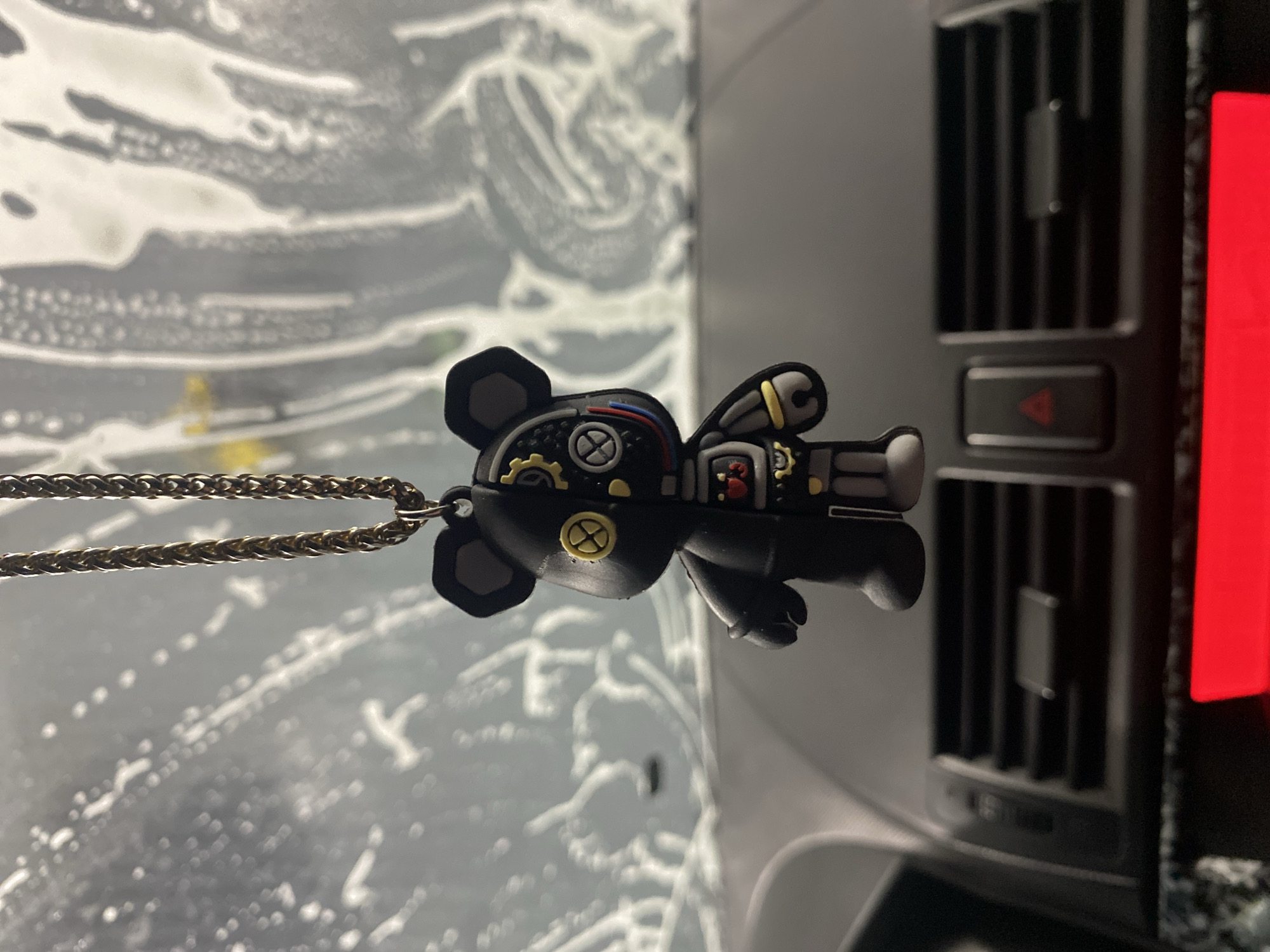 The image depicts a mechanical teddy bear pendant hanging from a gold chain. The bear is black with grey limbs, yellow and red details, and metallic accents suggesting a steampunk or robotic aesthetic. The pendant appears to be positioned inside a car, with a blurred view of what seems like a car wash or window covered in soapy water in the background. To the right of the pendant is a section of the car's dashboard including an air vent and hazard lights button. The red light of a car feature like a car radio or emergency lights feature is visible on the right. There is no evidence in the image that would allow a location in the city of Votkinsk, Russia to be confirmed.