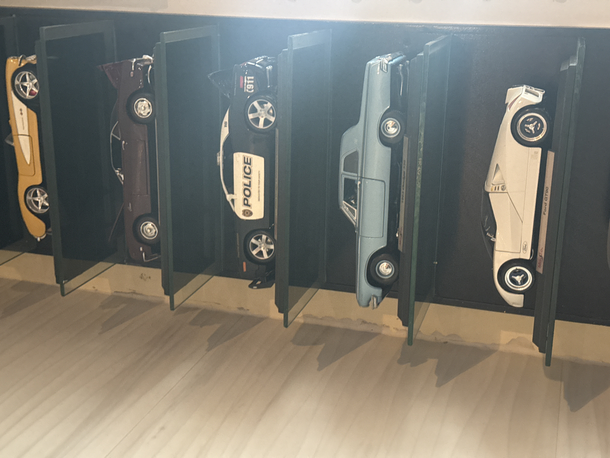 The image shows a collection of five die-cast model cars displayed in individual compartments within a dark enclosure. The cars are arranged in a row. From left to right, the models appear to be a gold sports car, a maroon classic car, a white police car with "POLICE" written on its side and the number 911 on top, a light blue car with visible window details, and a white Ford GT90 model. The vehicles are positioned vertically within their compartments, with their wheels facing the viewer. The floor beneath the display is a light, wooden surface. The lighting in the scene is somewhat dim, causing shadows to fall on the floor. There is no indication of the city being Puebla, Mexico or any evidence of people or interactions within the image.