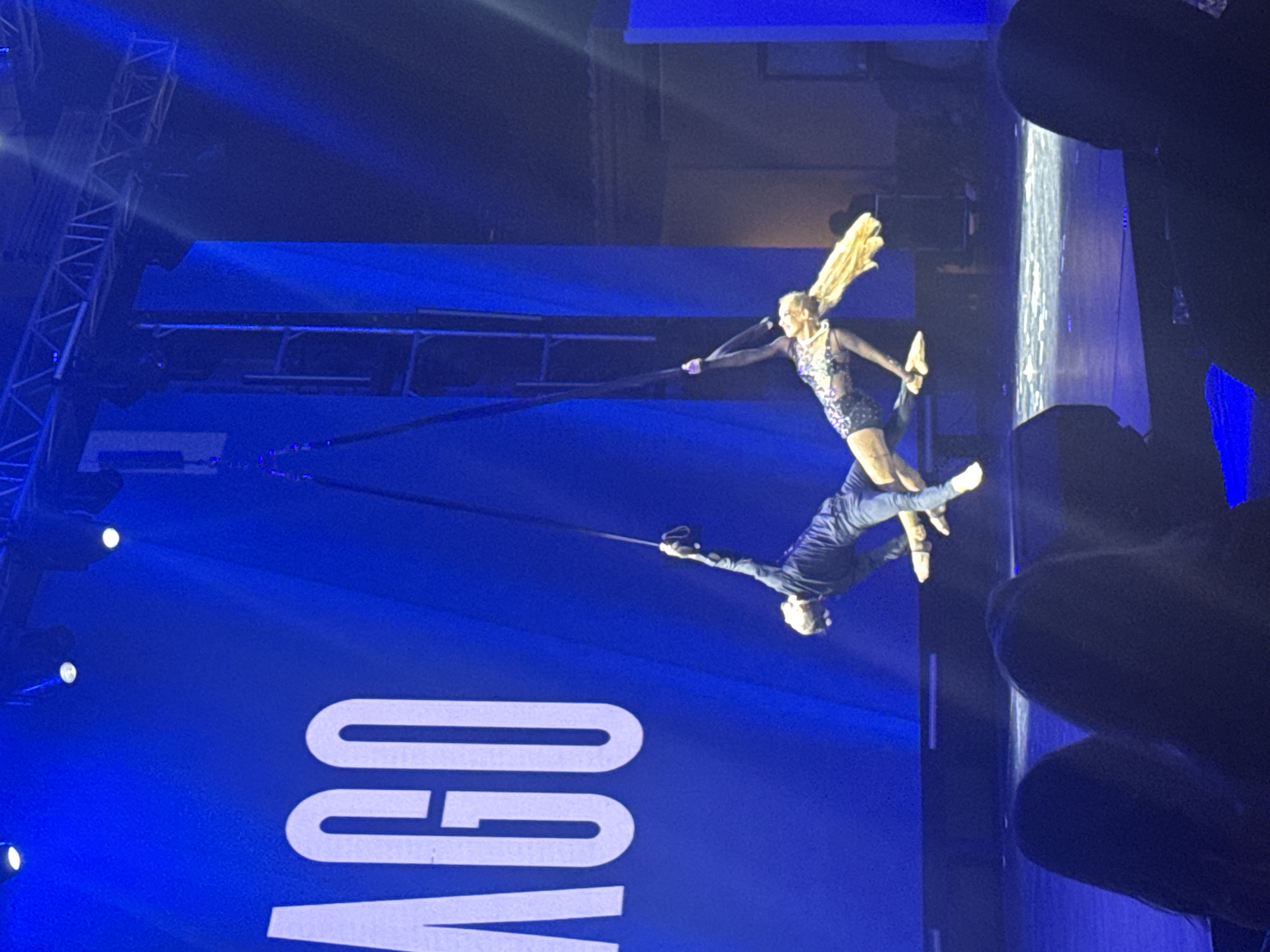 Two aerialists perform on silks in front of a large blue backdrop with the word "AGO" in white lettering. The aerialists are suspended in mid-air. One aerialist, wearing a dark long-sleeved shirt and jeans, is holding onto one silk with their left hand and has their legs extended. The other aerialist, wearing a sparkly black leotard and tights, is holding onto another silk with their left hand and is positioned below and to the right of the first aerialist. Their legs are extended and their blonde hair is flowing. The stage is lit with blue lights, and spotlights are visible from above. In the foreground, the silhouettes of an audience are visible. The location is identified as Manavgat, Turkey, suggesting this is a performance venue in that city.