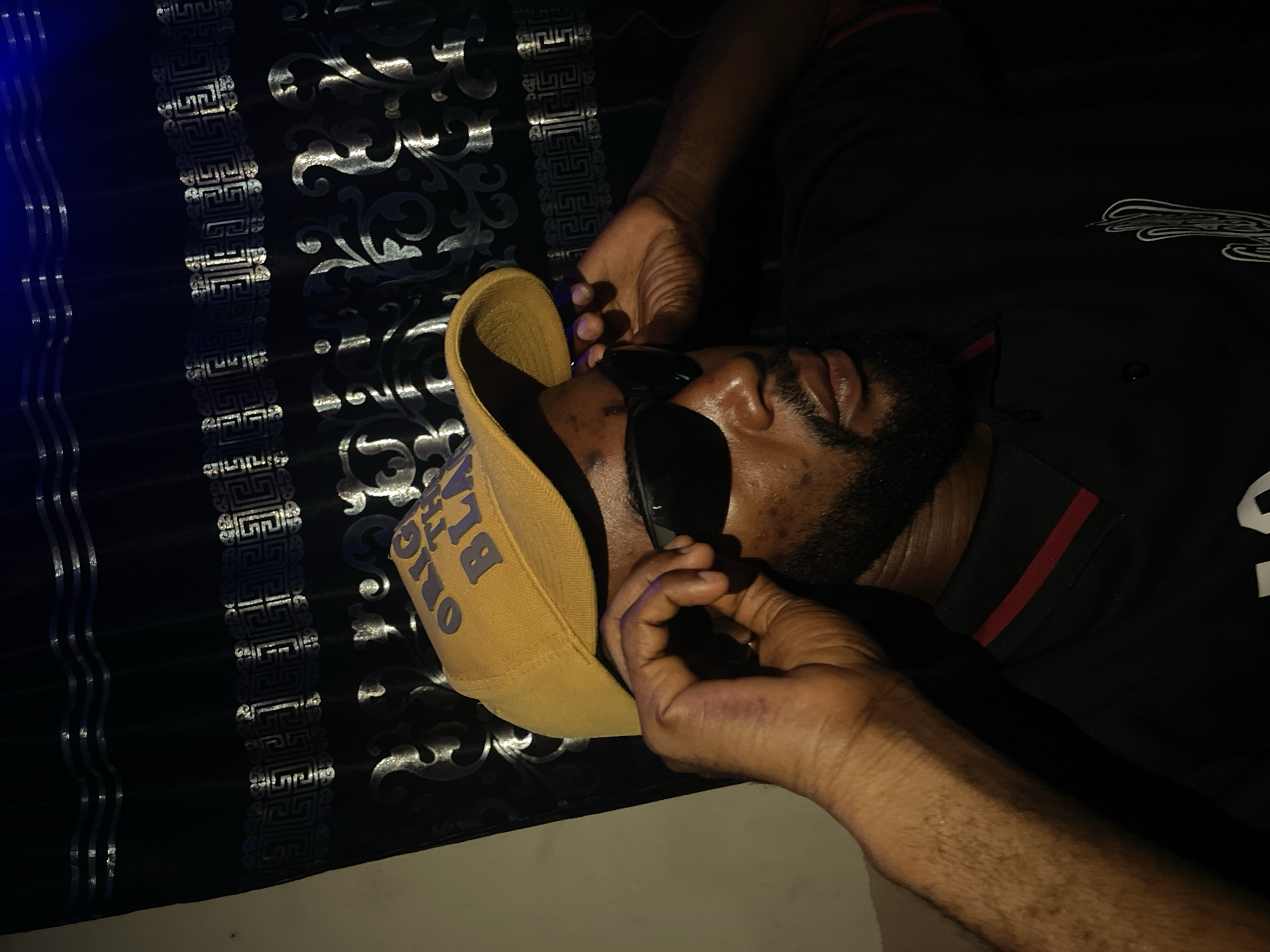 A man with dark skin and a beard is lying on a bed in an indoor location. He is wearing sunglasses and a tan-colored cap with blue lettering that reads "ORIGINAL THE BLACK". He is wearing a black shirt with a red stripe on the collar and a white design on the shoulder. His hands are holding the sunglasses. The bed has a black and silver patterned headboard. The location is unknown.