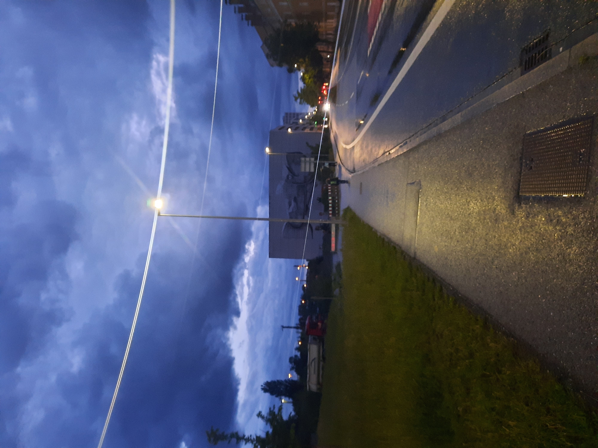 An external, eye-level, vertical shot captures a city street and sidewalk at dusk. The sky is filled with dark, dramatic clouds, suggesting an impending storm. A paved sidewalk, illuminated by a streetlamp, runs along the left side of the frame, bordered by a strip of green grass. The sidewalk transitions into a wet asphalt road to the right, with white lane markings visible. A streetlamp stands tall on the sidewalk, its light casting a warm glow on the wet pavement. In the distance, buildings are visible, including a large, modern structure with a mural on its side. A few vehicles, including a truck and cars, are present on the road, with their headlights on. Two figures are walking on the sidewalk in the mid-ground. The overall atmosphere is moody and somber due to the dark sky and wet conditions.