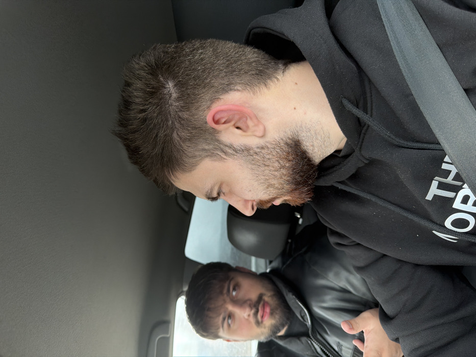 Two adult males are observed seated inside a vehicle. The scene is located in Gabarnitsa, Bulgaria.
The male in the foreground, positioned to the right, is seen in profile, facing left. He has short, dark brown hair and a reddish-brown beard. He is wearing a black hooded sweatshirt with a partial "THE NORTH FACE" logo in white visible on the chest. A grey seatbelt diagonally crosses his torso from his right shoulder to his left hip. His right ear exhibits a reddish hue. His gaze is directed towards the male in the background.
The second male, situated in the background to the left, is partially visible from the chest up, facing towards the right foreground male. He has dark, medium-length hair with bangs and a dark beard and mustache. He is dressed in a black, possibly leather or synthetic, puffer jacket. His right hand is raised, palm slightly upwards, indicating an active gesture, possibly during speech. His mouth is slightly open, suggesting he is speaking.
The interior of the vehicle features a dark grey headliner and black seat upholstery. The exterior environment visible through the windows is blurred, suggesting movement or a shallow depth of field, with an overcast sky.