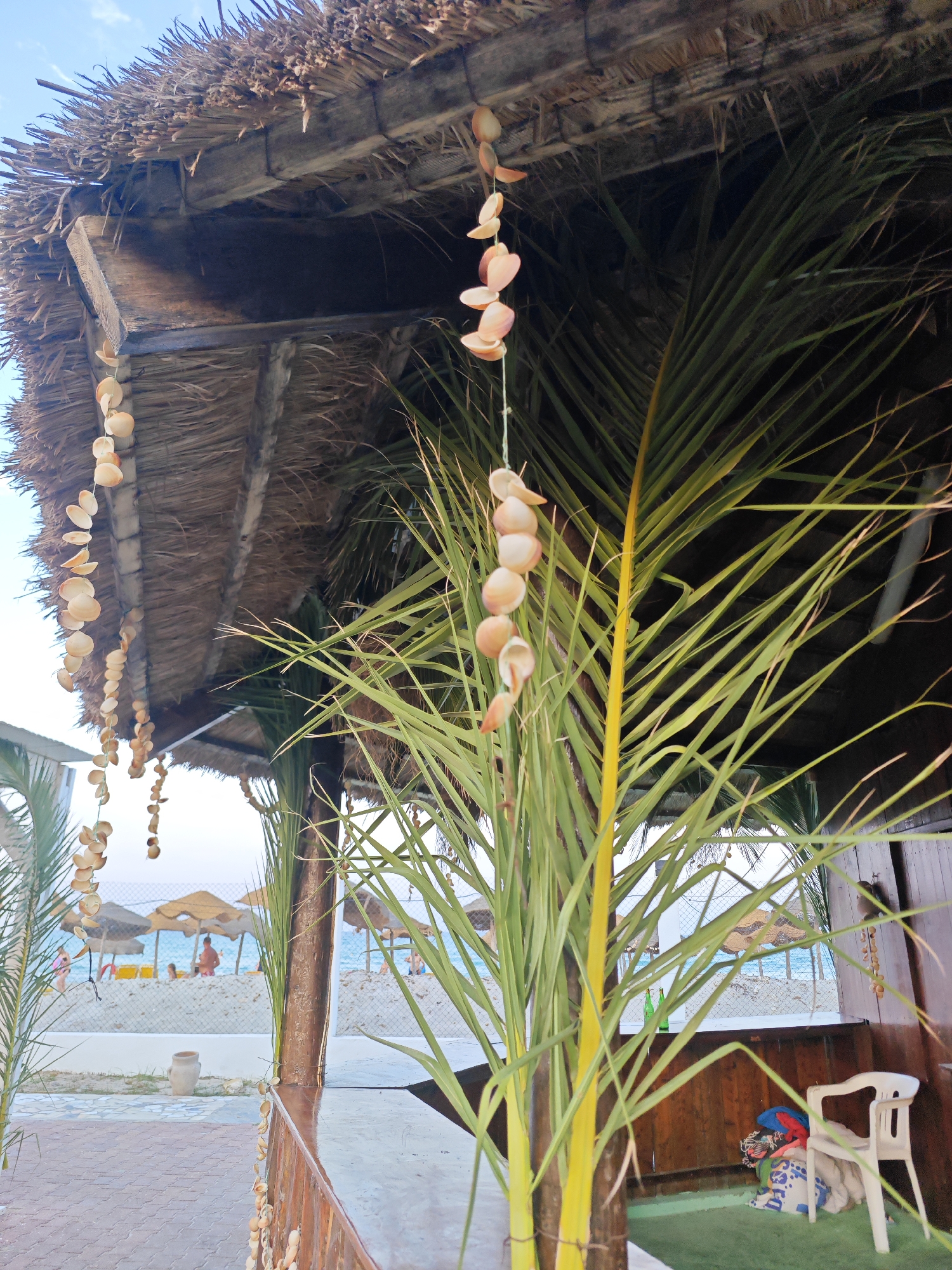 This image shows a detail of a thatched roof beach shelter with hanging seashell decorations and palm fronds. In the background, a beach with umbrellas and people is visible, suggesting a location in Mahdia, Tunisia. The foreground shows the rustic construction of the shelter, with wooden beams and straw thatch. A white plastic chair and some bags are placed in the shelter. The overall impression is one of a relaxed, tropical beach setting.