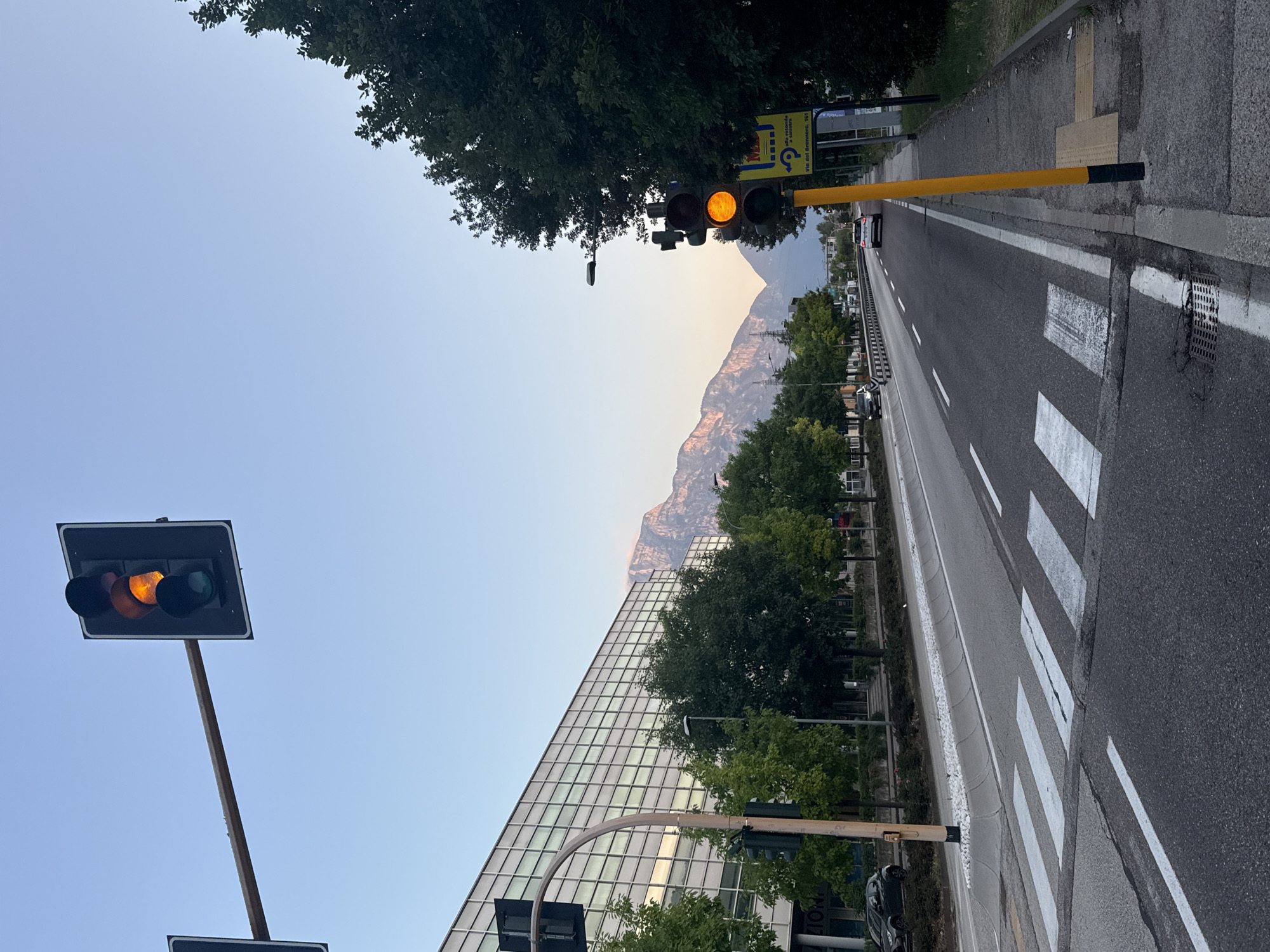 The image shows a traffic light at an intersection in Trento, Italy. The traffic light is orange, indicating that the signal is about to change. In the background, there is a modern building with a glass facade, trees, and mountains. A pedestrian crossing is visible in the foreground.