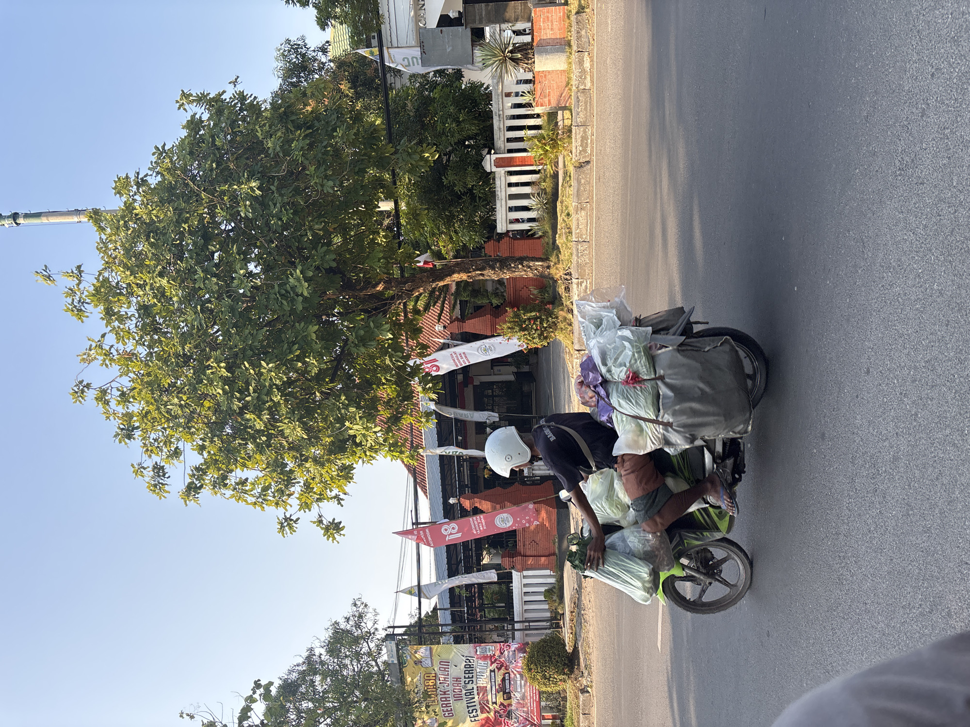 A man wearing a white helmet and black t-shirt rides a green motorcycle laden with large bundles of produce wrapped in plastic. He wears brown shorts and blue flip-flops. The motorcycle has two large gray saddlebags filled with more produce. In the background, a large tree with green leaves is prominent. Behind the tree, a building with red brick accents and a white fence is visible, along with several flags and banners. One banner on the left has text in Indonesian, including "GERAK JALAN INDAH FESTIVAL SERABI." A tall, slender tower, possibly a minaret, is visible in the far distance to the right. The scene takes place on a paved road, suggesting an urban or suburban environment. The lighting indicates it is daytime, possibly late afternoon, given the angle of the shadows. The overall impression is of a vendor transporting goods in Cirebon, Indonesia.
