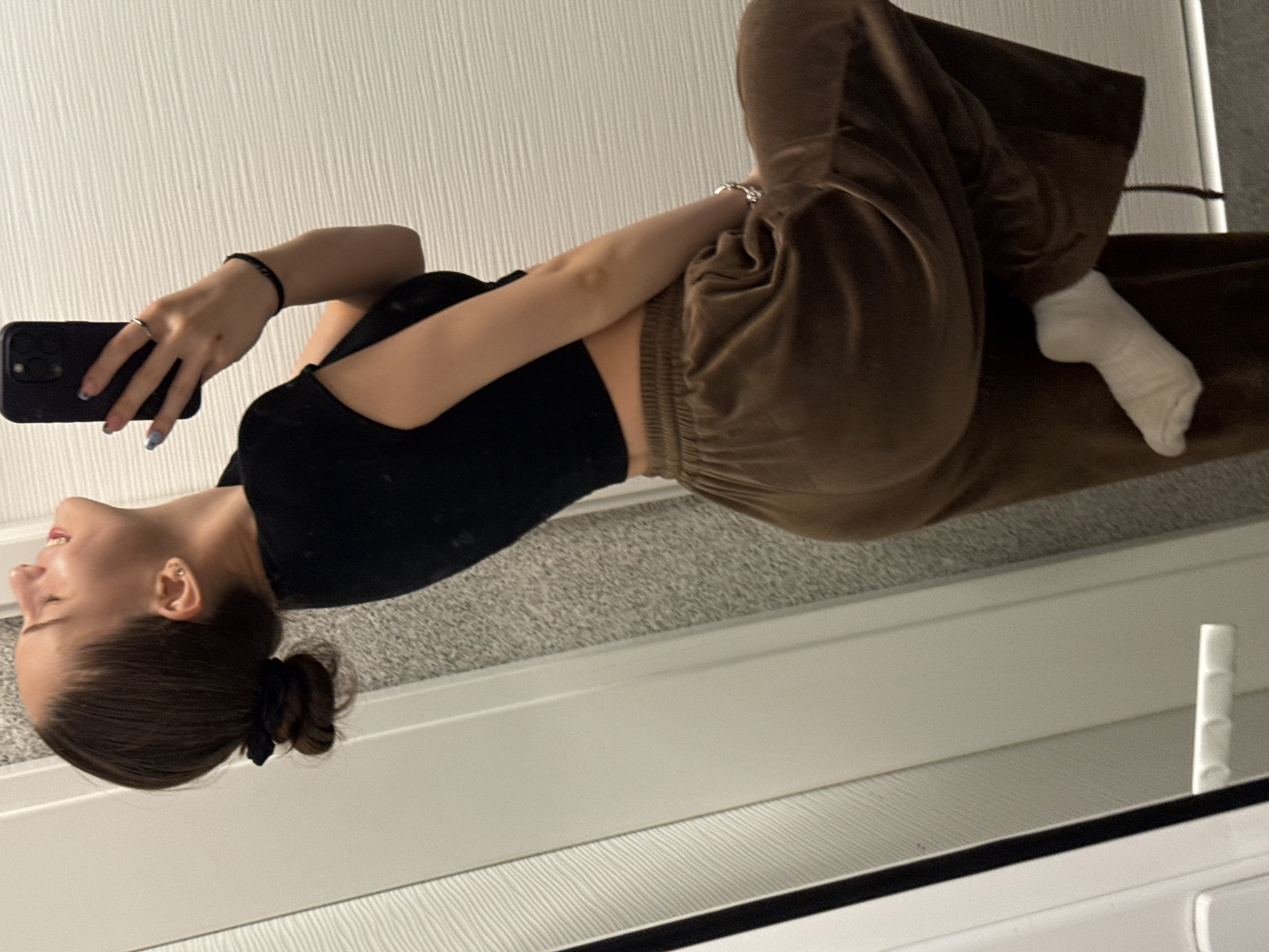 A young woman is depicted in a mirror selfie. She is wearing a black crop top and brown velour pants, with a white sock on one foot. She is holding a black smartphone, presumably taking the picture. Her hair is tied back in a bun. The background consists of a white wall with a textured surface and a grey carpet. The reflection suggests an indoor setting, likely a home interior. There is no information available to identify a specific location in Sofia, Bulgaria.