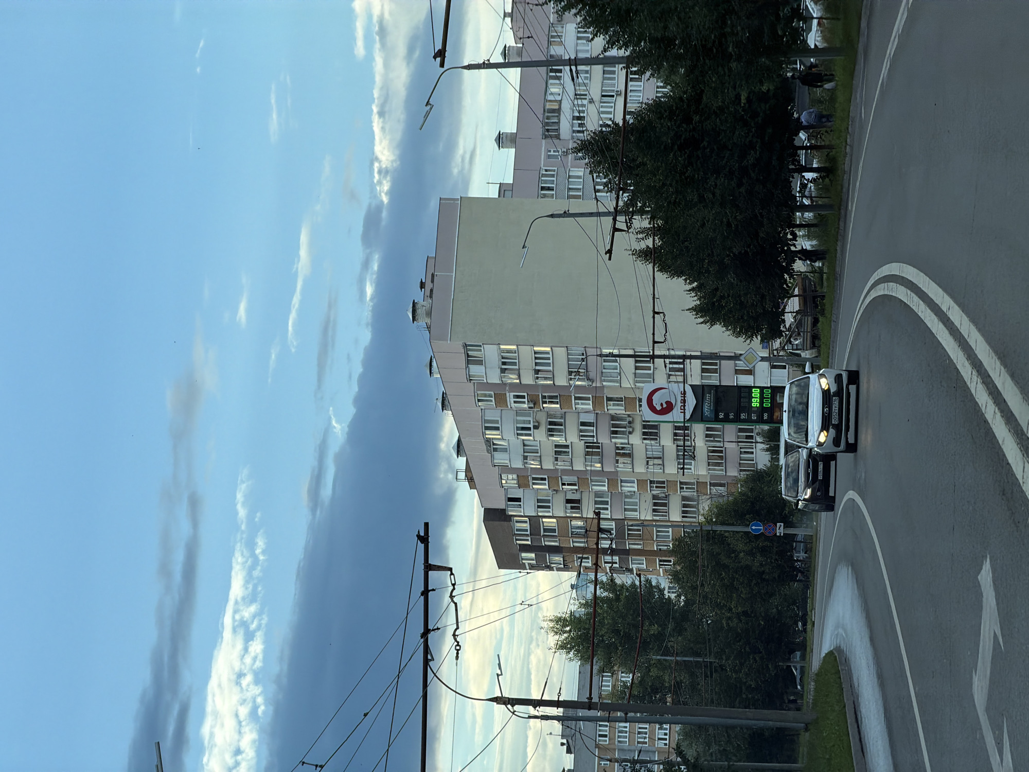 This image depicts a street scene in Kazan, Russia, under a partially cloudy sky. A multi-story apartment building with numerous windows is prominently featured on the right side of the frame. In the foreground, a gray road curves to the left, with white lane markings. Two cars are visible on the road: a black car in the left lane and a white car in the right lane. The white car has its headlights on. Overhead, trolleybus wires crisscross the sky, supported by poles. Trees line the right side of the road, behind which the apartment building stands. A gas station sign with prices displayed in green digits is visible on the right side of the image, with "IBIS" and "XTREM" text on it. There are several people walking on the sidewalk on the far right, and a few birds are perched on the trolleybus wires and in the sky. The overall impression is a typical urban street during daylight, possibly late afternoon or early evening, given the quality of light.