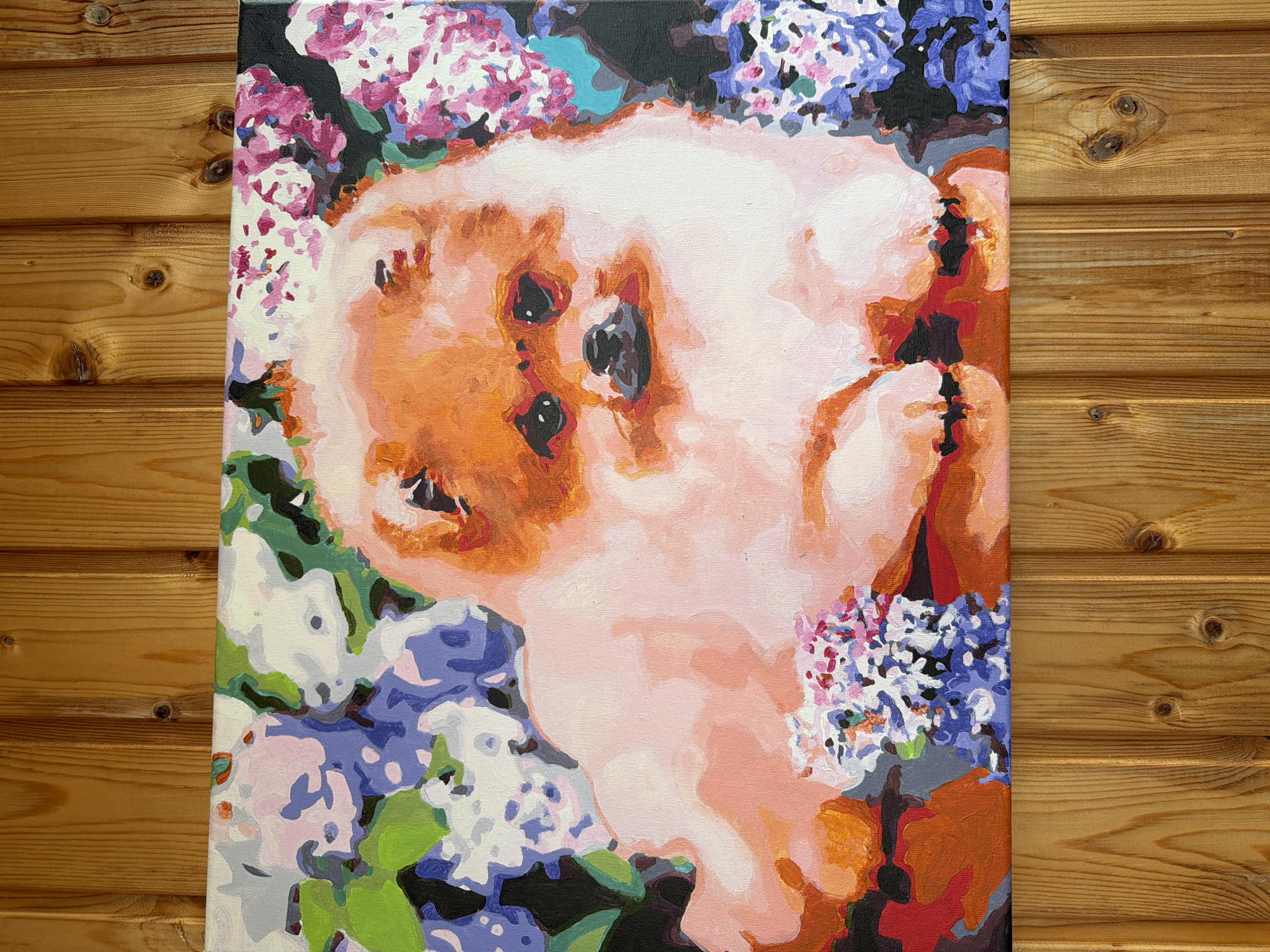 The image shows a painting leaning against a wooden wall composed of horizontal planks. The painting is a portrait of a small dog, possibly a Pomeranian, surrounded by pastel-colored flowers, likely lilacs. The dog is rendered in broad, visible brushstrokes with colors of pink, orange, and white, with black eyes and nose. The flowers are purple, pink, and white with hints of green foliage. The background of the painting is also filled with flowers and dark areas. The painting appears to be oil or acrylic on canvas. The location is unknown.