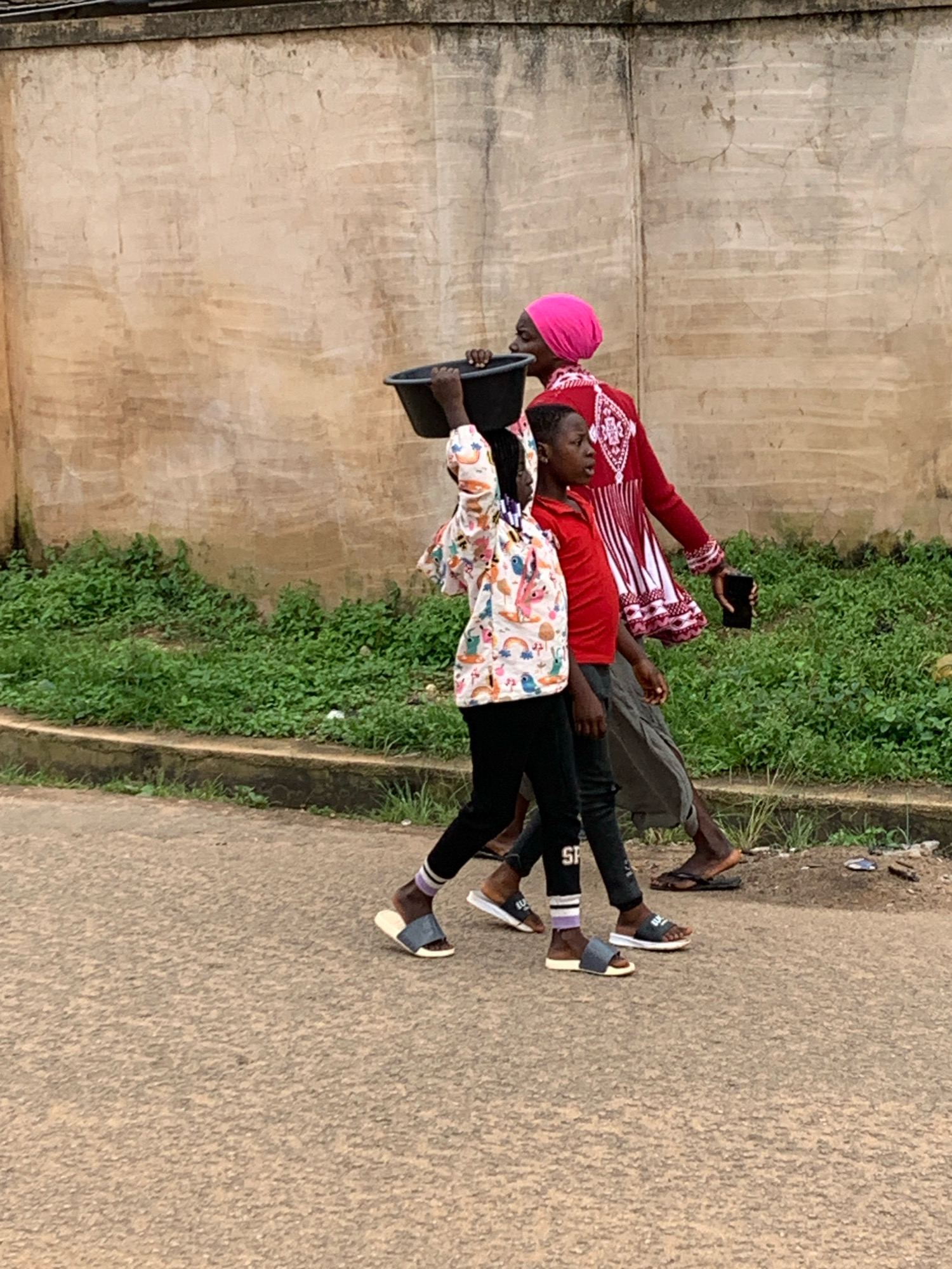 In Jos, Nigeria, two individuals are walking on a paved road beside a wall covered in patches and a strip of dense green vegetation. The person in front, a child, is carrying a large black basin on their head. They are wearing a colorful long-sleeved shirt with a pattern of cartoon characters and rainbows, black pants, and sandals. To their right, an adult walks, dressed in a bright red garment with white patterns and a pink head covering. They are holding a black mobile phone in their right hand. The ground beneath them is a textured asphalt surface.