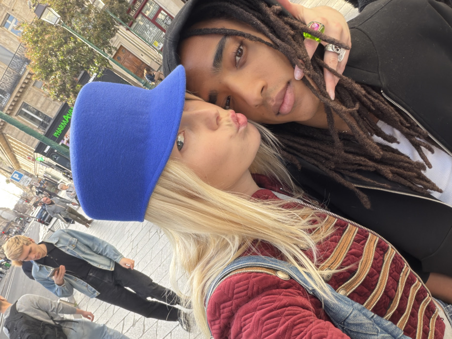 A close-up selfie captures two individuals outdoors on a sunlit street in Paris, France. The primary subject, a blonde-haired female, wears a distinctive royal blue felt hat with a flat top and short brim, a dark red quilted jacket with horizontal gold-colored stripes and metallic buttons, and an underlying denim garment. She looks directly at the camera with her lips pursed in a kissing gesture. Her left hand, adorned with light pink nail polish and multiple rings (including one with a prominent green and pink gemstone), rests on the shoulder of the second individual. This second individual is a dark-skinned male with long, dark dreadlocks, wearing a black hooded zippered jacket over a white ribbed tank top. He also looks towards the camera with a neutral expression.
The background shows a typical Parisian street scene. The ground is paved with light-colored cobblestones or pavers. Multi-story buildings with classic Haussmannian architecture, featuring light-colored stone facades, ornate windows, and balconies, line the street. Identifiable storefronts include a "PHARMACIE" marked by a green cross sign and a partially visible "BIJOUTERIE" sign. A blue parking sign with a white "P" and the word "Payant" is visible. Several pedestrians are present in the mid-ground, notably a blonde-haired male in a light blue denim jacket, black polo shirt, and black pants, who is walking away from the camera while engaged with a smartphone and wired headphones. A traditional black street lamp and a tree with green leaves are also visible. The scene is illuminated by bright daylight.
