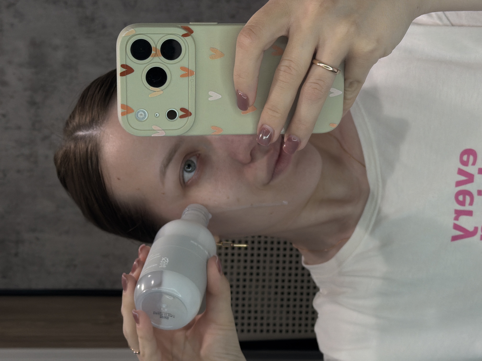 A woman is taking a selfie in an indoor setting, likely a bathroom, as suggested by the mirror reflecting a tiled wall behind her. She is holding a phone with a light green case decorated with small hearts in her right hand, which is raised to obscure part of her face. The phone has a triple camera setup. She wears a gold ring on her right ring finger, and her nails are painted with a mauve color. She is applying a skincare product from a grey and white bottle held in her left hand onto her cheek. A small streak of white product is visible on her skin. She wears a white t-shirt with pink lettering on it. The background includes a section of wall with a dark, horizontal division. The scene is well-lit. There is no evidence of Reshetnikovo, Russia.