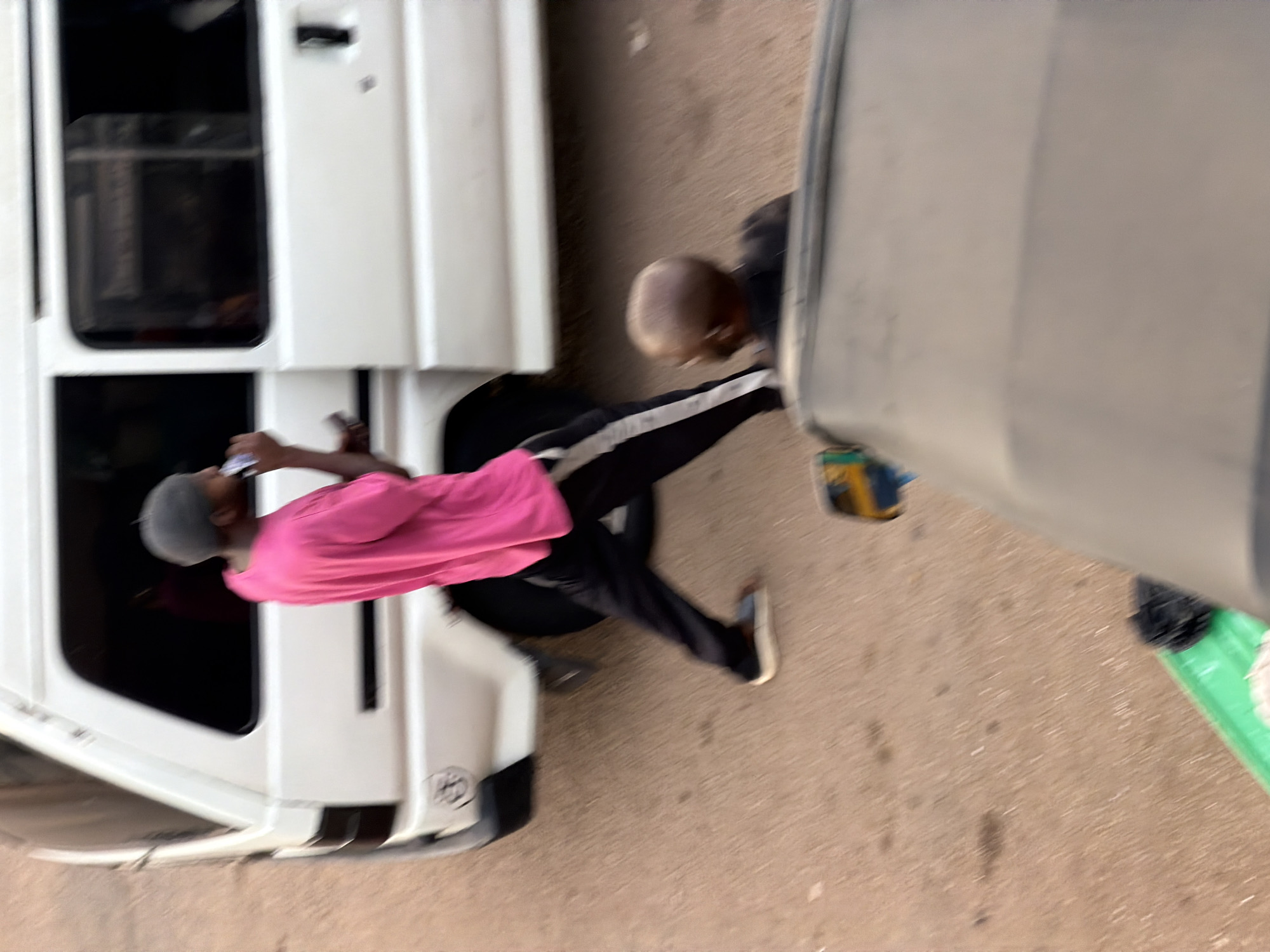 In Bukuru, Nigeria, a person wearing a gray beanie and a bright pink t-shirt is standing next to a white van, holding a phone to their ear. They are wearing black pants with white stripes and dark shoes. The van has tinted windows and a handle on the side. Further back, another person with a shaved head and wearing a black shirt is visible, partially obscured by a gray vehicle in the foreground. The scene appears to be on a street with a paved surface, and a small green object is visible in the lower left corner.