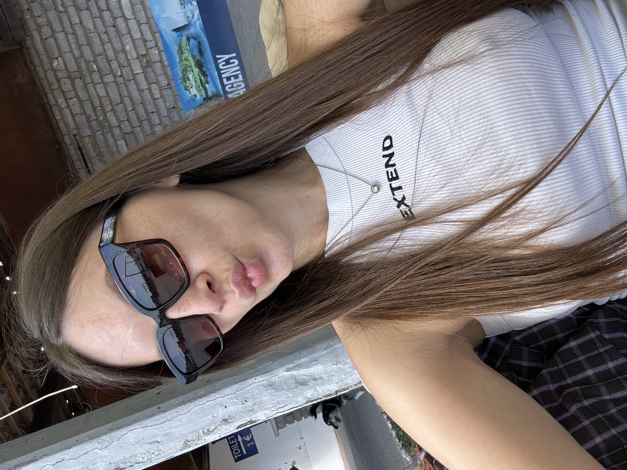 A young woman poses for a photo, wearing sunglasses and a white ribbed tank top with the word "EXTEND" printed on it. She has long brown hair and a necklace. The scene appears to be outdoors in Budva, Montenegro, as evidenced by the "AGENCY" sign referencing local tourism and the visible street and building details. A blue and white sign indicating "TOILET 1€" is also visible. The woman is wearing a plaid skirt or shorts.