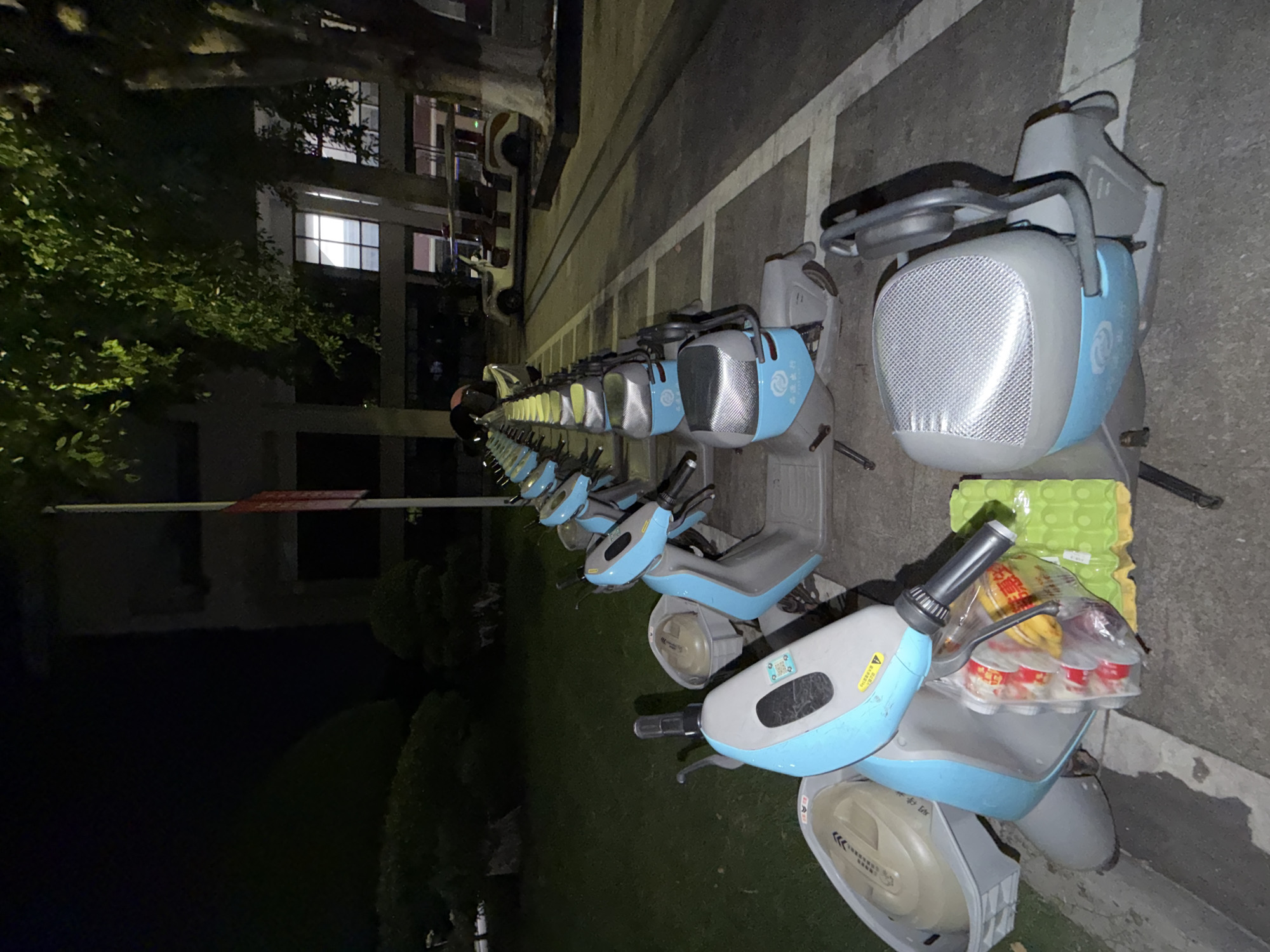 An outdoor scene at night displays a row of parked, light blue and gray electric scooters. The scooters are lined up on a paved surface marked with white lines, suggesting a designated parking area. In the foreground, one scooter is most prominent, with a plastic basket containing groceries and a green egg carton beside it. The background shows a building with illuminated windows, trees with green foliage, and a small, white electric vehicle. The overall setting appears to be a public space in the city of Jiangjin, China.