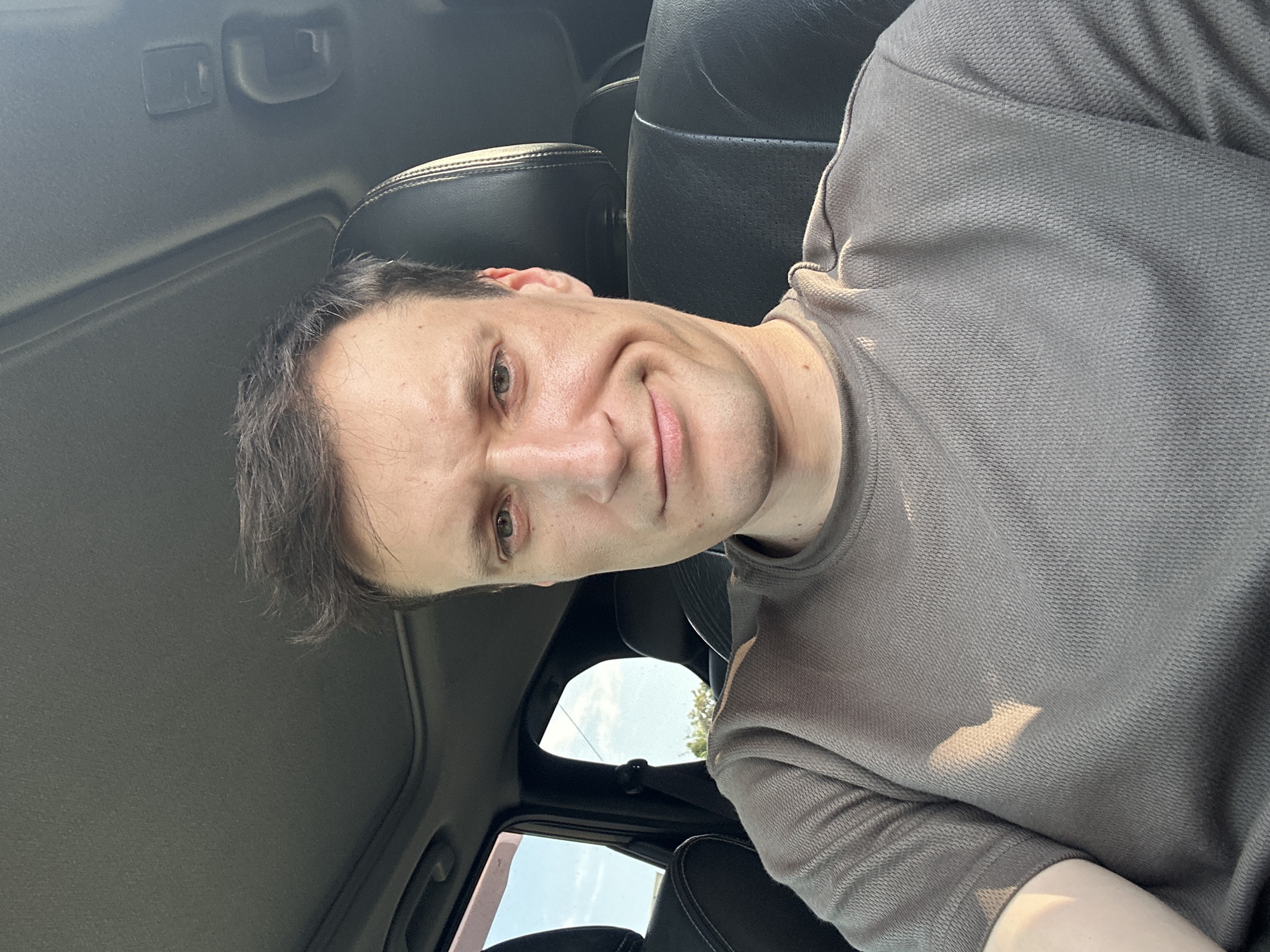 A man is seated in the driver's seat of a vehicle, looking directly at the camera. He has short, messy brown hair and is wearing a gray t-shirt. The interior of the car is visible, including the black leather headrest and part of the sunroof. The sun is shining, casting a shadow on the man's left shoulder. The scene appears to be set in Shatava, Ukraine, based on the provided context.
