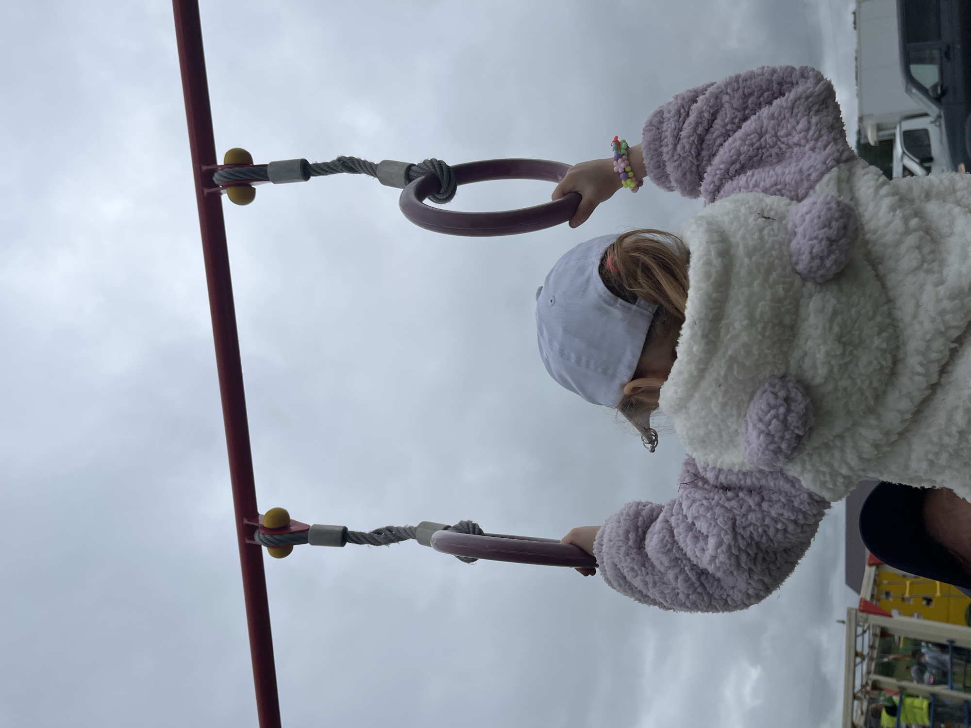 An overhead perspective captures the back of a child holding two dark purple gymnastic rings suspended from a red horizontal bar under an overcast, light gray sky in an outdoor recreational area in Unknown, Unknown. The child wears a white baseball cap, a white fluffy jacket with lavender sleeves and ear-like details, and a colorful, flower-shaped beaded bracelet on the right wrist. Each gray braided cable supporting a ring is attached to the red bar via a metal bracket with yellow stoppers. In the blurry background on the left, several individuals, some wearing high-visibility vests, are visible near playground-like structures and scaffolding. On the right, parked vehicles, including a white pickup truck and a dark SUV, are partially visible in front of a white building.