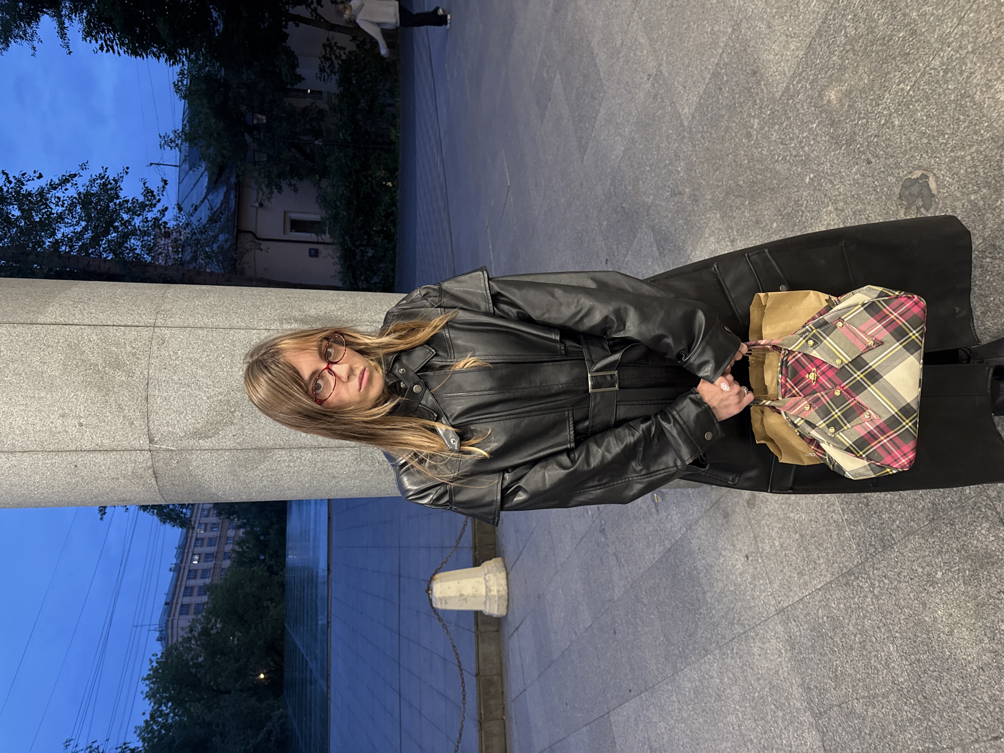 A young woman stands in front of a large concrete column, holding a patterned handbag. She is wearing a long black leather coat and red-rimmed glasses. The background shows a paved area with some trees and buildings, indicating an outdoor urban environment, possibly in Moscow, Russia given the architectural style. The lighting suggests it is dusk or early evening.
