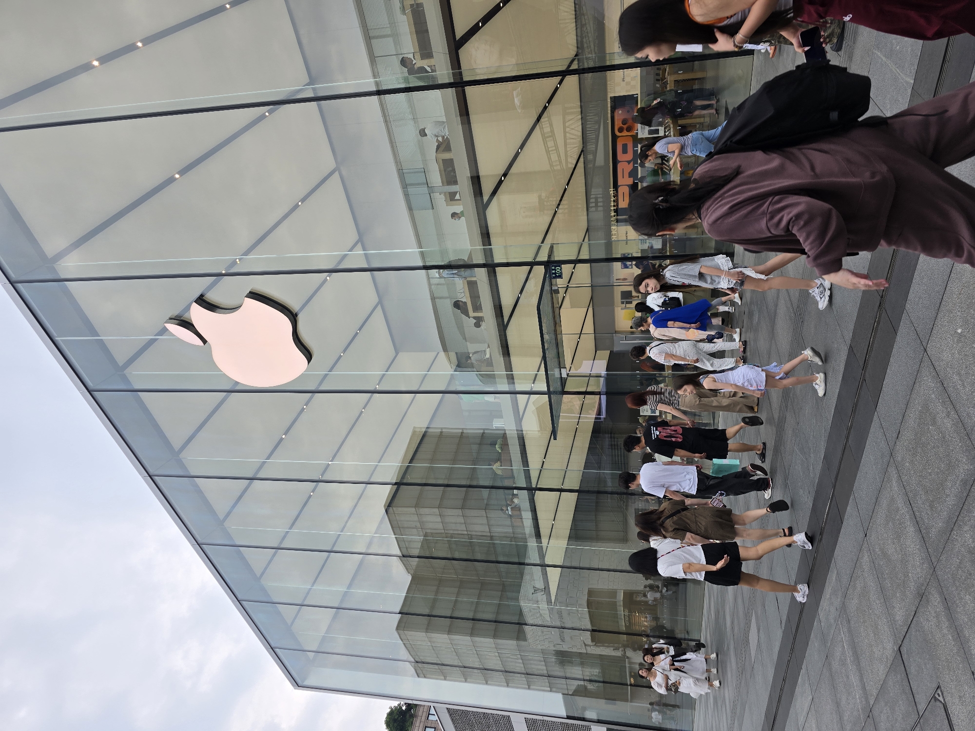This image captures the exterior of a modern Apple Store, characterized by its large, transparent glass facade. The iconic Apple logo, rendered in a light, metallic hue, is prominently displayed on the upper left side of the glass. The building's sleek, angular design features multiple glass panels and metal frames.
Numerous people are visible in the scene. Outside the store, a crowd walks along a paved sidewalk, some appearing to be queuing or heading towards the entrance. They are dressed in casual modern attire. One person in the foreground wears a dark brown tracksuit and carries a black backpack. Other individuals wear t-shirts, shorts, dresses, and sneakers. Inside the store, reflected through the glass, more people can be seen interacting with products and staff, typical of a retail environment. The reflections also show parts of the store's interior, including display tables and overhead lighting.
The sky, visible in the upper left, is overcast, suggesting a cloudy day. The overall lighting indicates daytime, likely mid-day or afternoon, without harsh shadows. The ground is a uniform grey pavement. The store's modern architecture and the presence of many people suggest a bustling urban environment, consistent with a shopping district in Shangcheng District, China.
Visible text includes the prominent Apple logo. Reflected on the glass, partially obscured, are words that appear to be "PRO" and possibly "iPhone", along with a numerical sign that reads "100".