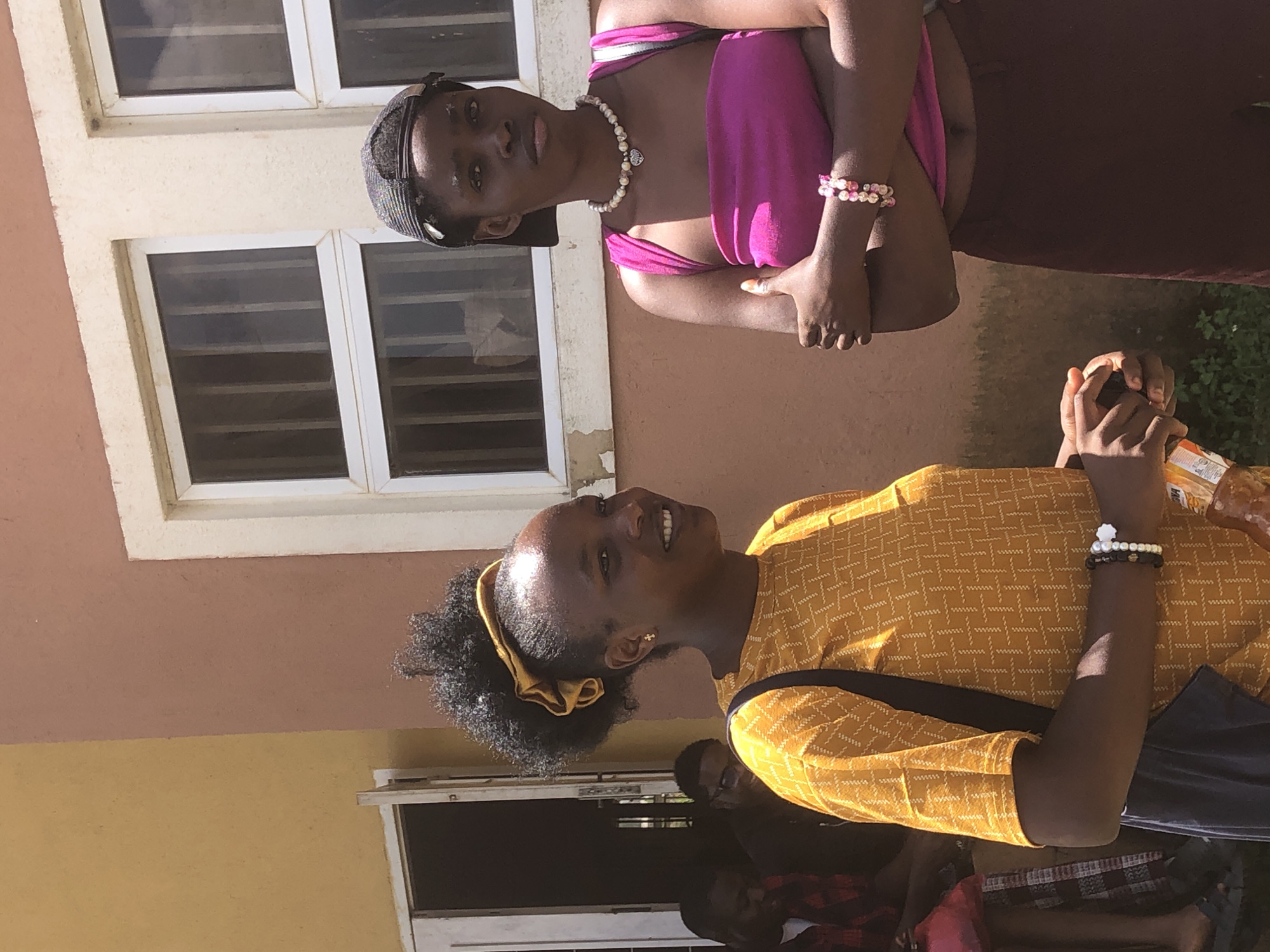 Two young Black women are standing outside a building with a pale yellow and terracotta-colored facade. The woman on the left, with her hair pulled up in a high ponytail with a yellow scrunchie, wears a yellow patterned long-sleeved shirt and a dark shoulder bag. She is smiling and looking to her right. The woman on the right wears a bright pink strapless top, dark pants, a black baseball cap, and a pearl necklace with a heart pendant. She has her arms crossed and is looking forward. Behind them, a doorway is open, revealing the interior of the building, and two men are partially visible. The environment suggests a sunny day. The building appears to be located in the city of Gwafan, Nigeria.