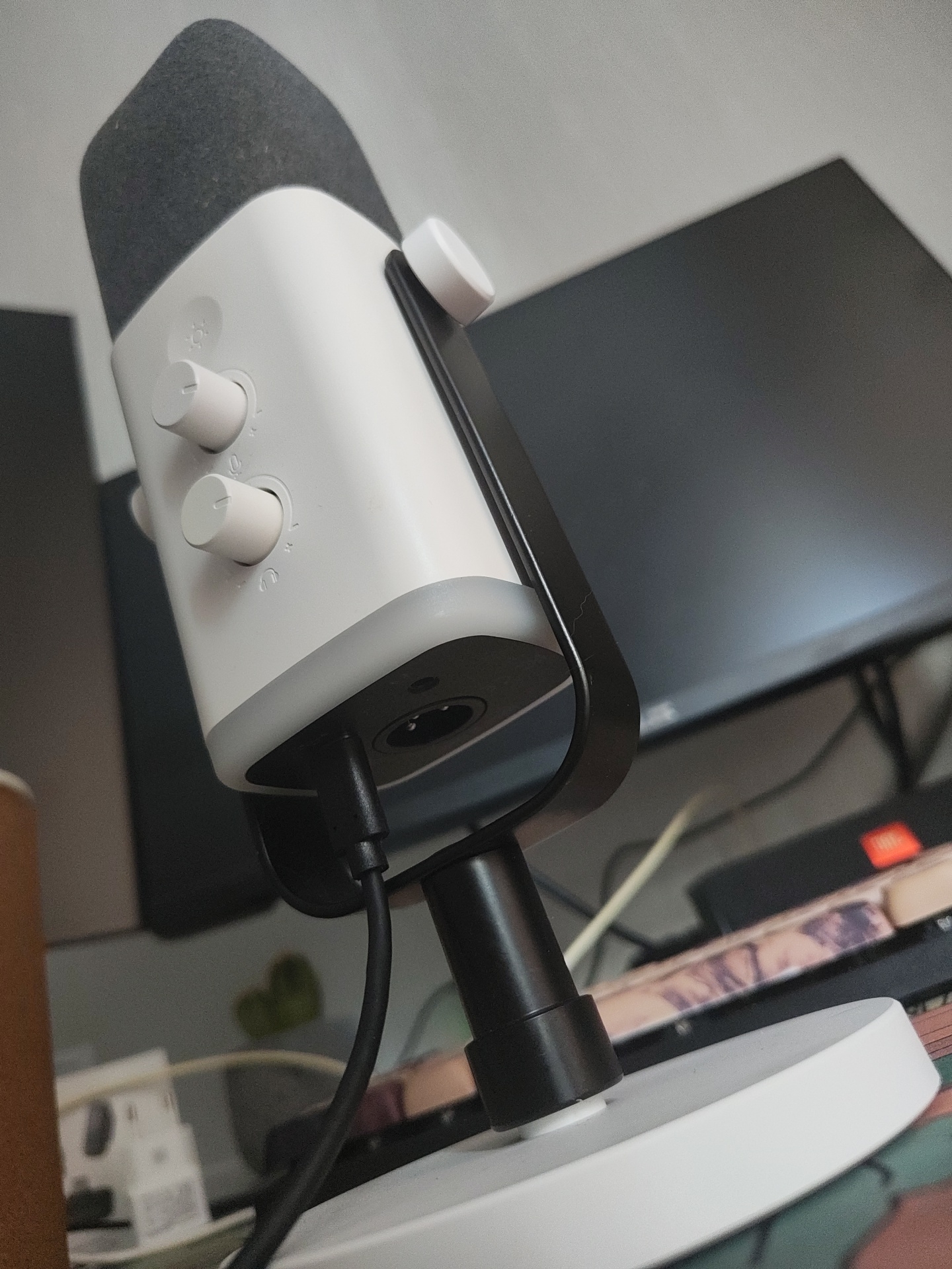The image shows a white and gray USB microphone on a desk. The microphone has two white knobs on its body, and a mesh-covered top. A black cable is plugged into the bottom of the microphone. In the background, a computer monitor is visible, along with other desk accessories and cables. The scene appears to be an indoor workspace. There is no indication of the city of Il’inskiy, Russia.