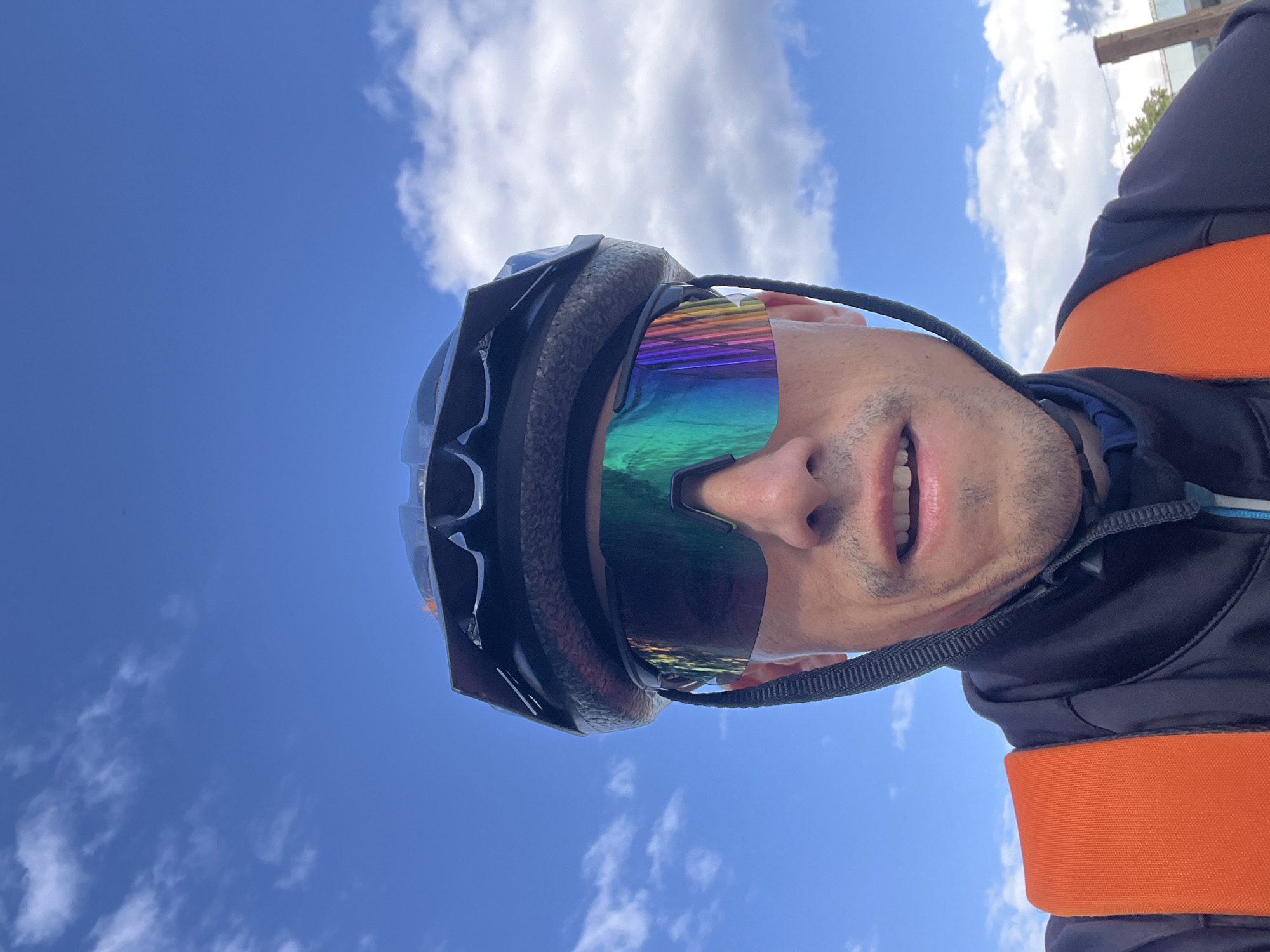 A close-up, dynamically angled shot captures a person wearing cycling gear against a bright blue sky with scattered white clouds, set in Chaikovsky, Russia. The image is vertically oriented, giving the impression the subject is looking upwards at the sky.
The central subject is a person, likely male, whose face is partially visible. They are wearing a dark grey or black cycling helmet. Dominating the upper face are large, wraparound sports sunglasses with highly reflective, iridescent lenses that shimmer with vibrant green, blue, purple, and yellow hues, reflecting the surroundings. A thin black strap, possibly from the helmet or glasses, extends along the side of their face.
Below the sunglasses, the person's nose and part of their mouth are visible, with lips slightly parted in a subtle smile, revealing white teeth. Light stubble is visible on their chin and upper lip. They are dressed in a dark-colored jacket, and a prominent, bright orange strap or band crosses their torso, suggesting a part of a backpack or specialized cycling attire.
The background is a clear, expansive blue sky dotted with fluffy white cumulus clouds, indicating bright daylight conditions, likely midday or early afternoon. The low perspective of the shot emphasizes the sky. The overall scene suggests outdoor athletic activity, most probably cycling, on a sunny day. No visible text is present.