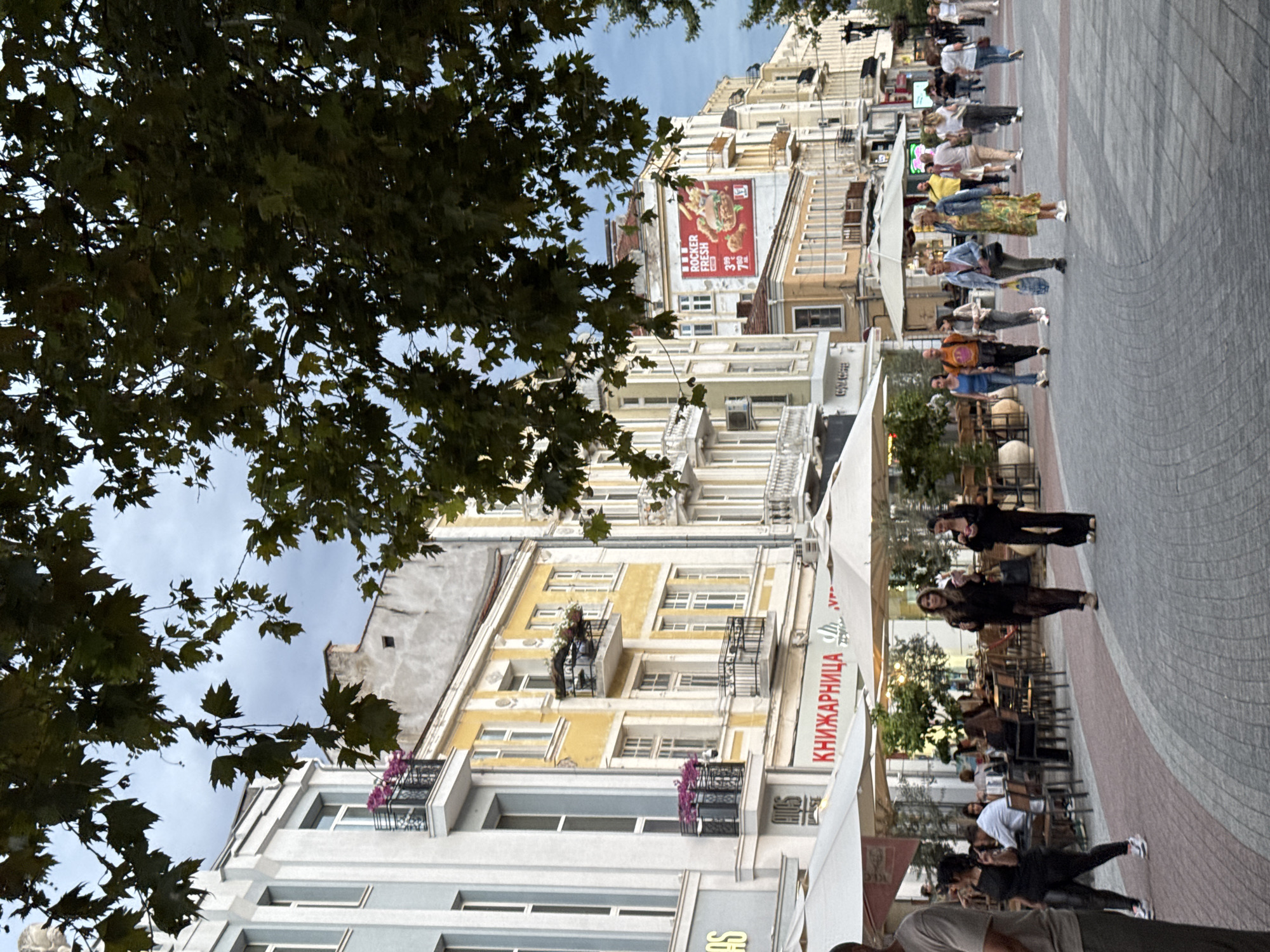 The image depicts a lively street scene in Plovdiv, Bulgaria, likely on the pedestrianized main street, known as "Glavnata." The perspective is from street level, looking down a wide, paved walkway lined with buildings on both sides.
In the foreground, several people are walking or gathered. On the left, a man in a gray t-shirt is partially visible. Further down, a woman in black leggings and a black top walks towards the viewer, with another woman in a black lace dress and black pants walking away. Behind them, several tables and chairs of outdoor cafes are visible under white umbrellas, with patrons seated and interacting.
The buildings lining the street exhibit a mix of architectural styles, common in Plovdiv's historical center. On the left, a light blue building with white trim and balconies is visible. Adjacent to it, a yellow building with ornate balconies and windows stands out. Further down the street, other buildings in similar styles, some with signs and advertisements, form a dense urban backdrop. A prominent billboard for "ROCKER FRESH" featuring a burger is visible on one of the buildings.
Trees with dense green foliage overhang the street from the top of the frame, casting shadows and adding a natural element to the urban environment. The sky above is overcast, suggesting a cloudy day. The overall impression is of a bustling city center with outdoor dining, shopping, and pedestrian activity.