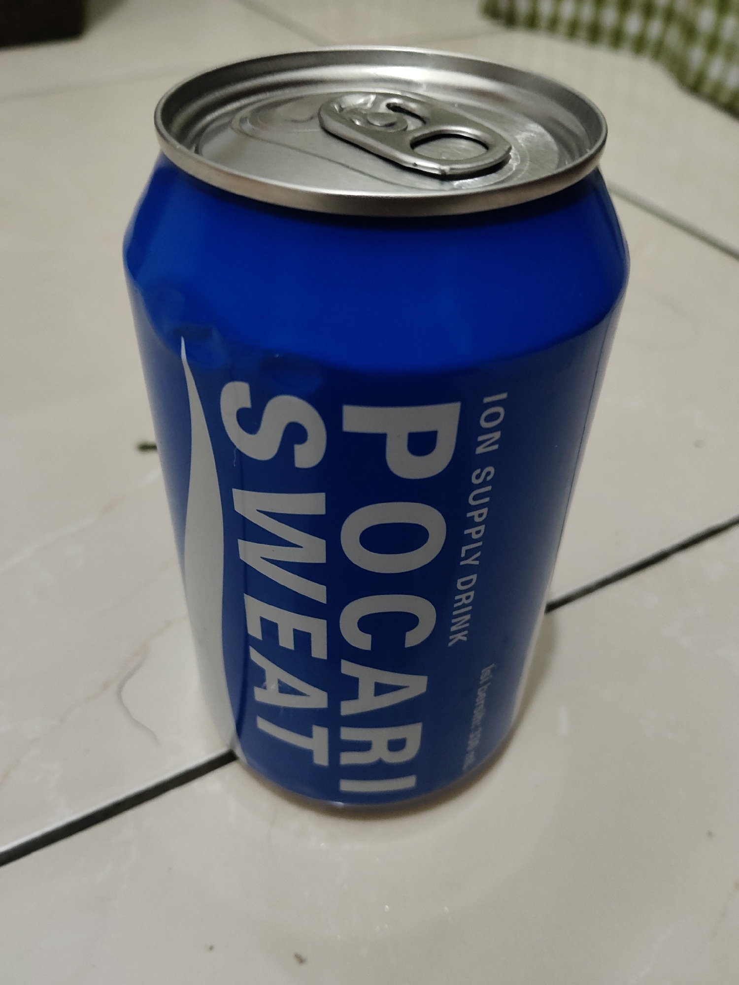 A blue can of Pocari Sweat is placed on a tiled surface. The can has white lettering that reads "POCARI SWEAT" vertically. The top of the can, which has a silver pull-tab, is visible. The background is out of focus, showing a checkered pattern and a glimpse of a white surface. The lighting suggests an indoor setting. The image does not contain any information that would allow for the determination of its location in Mekarmukti, Indonesia.