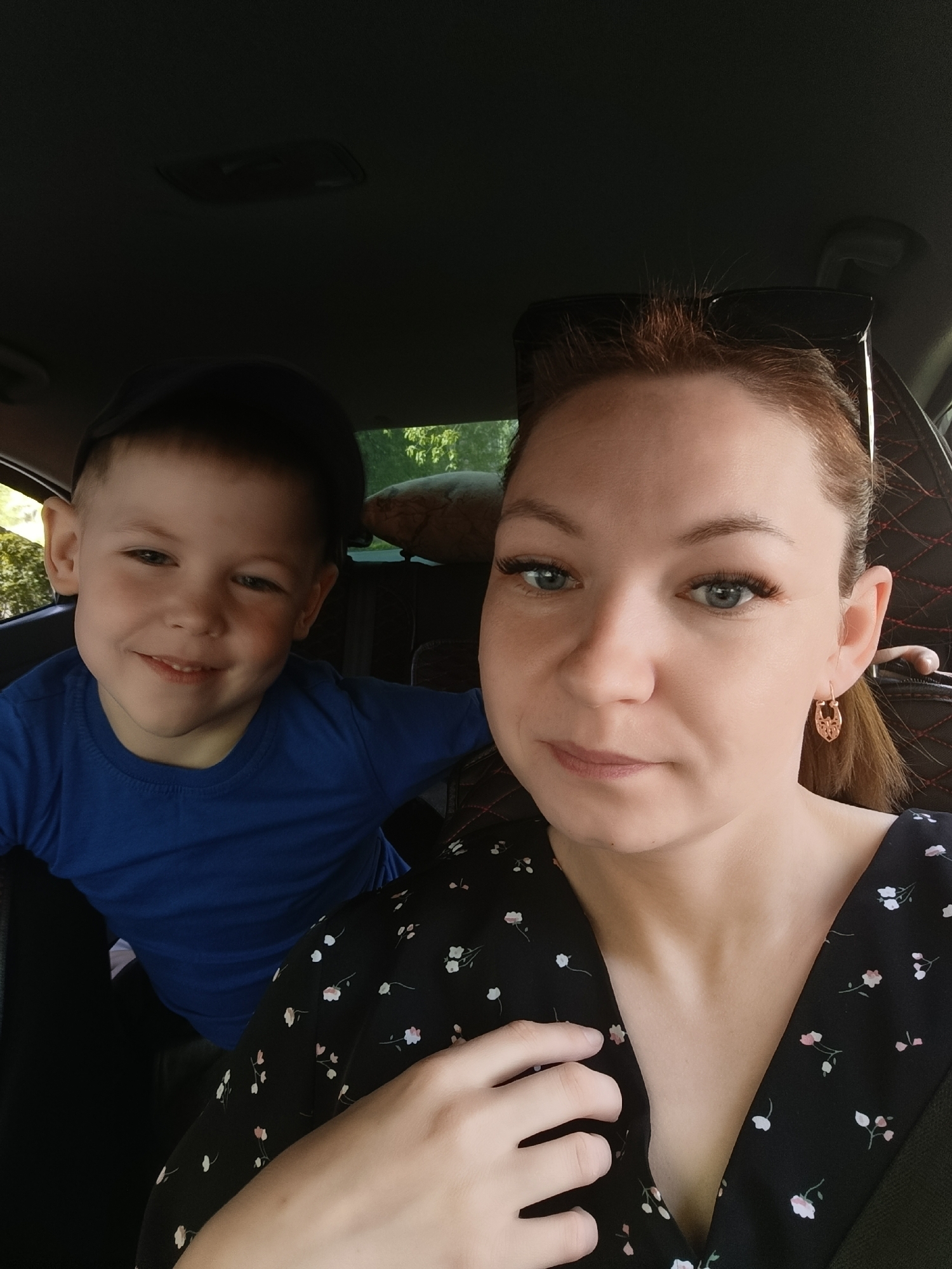 A selfie taken inside a car in Chaikovsky, Russia, shows a woman with fair skin, reddish-brown hair pulled back, and wearing a black floral print dress. She is looking at the camera. To her left, a young boy wearing a blue t-shirt and a dark cap is smiling. The car's interior is visible, including the ceiling, seats, and window with a view of trees outside.