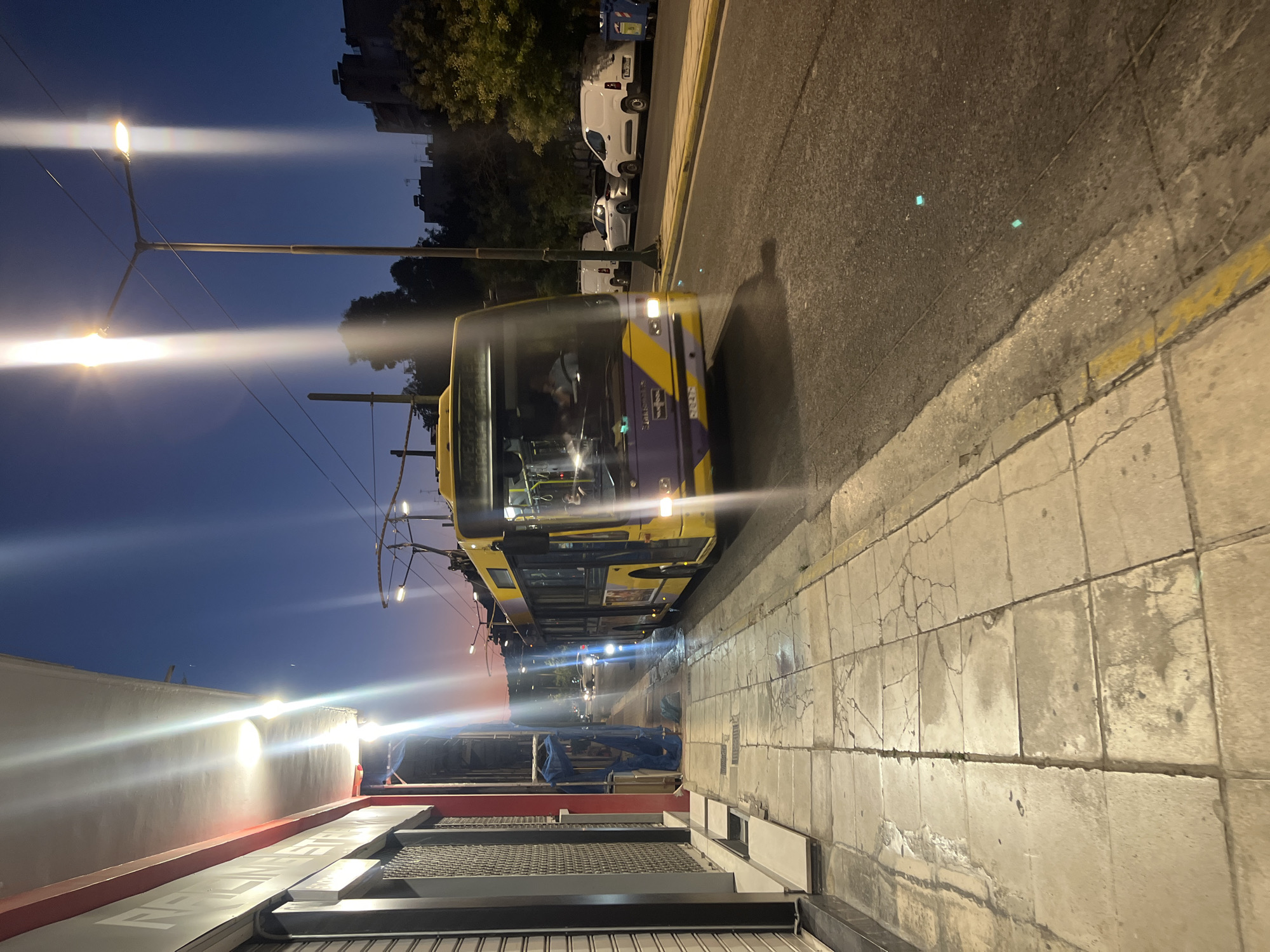 A yellow and purple trolleybus with its pantograph extended is traveling on the right side of a two-lane road. The trolleybus is illuminated by streetlights and its own headlights. It is nighttime, and the sky is a deep blue with a hint of orange near the horizon. The trolleybus is in motion, and its front is angled slightly towards the camera.
To the left of the road is a cracked concrete sidewalk. Along the sidewalk, there is a building with a red trim and closed metal shutters. The building appears to be commercial. Further down the sidewalk, there are some blue tarps covering an object.
On the right side of the road, there are several vehicles parked. One is a white van, and another is a silver car. There are also some trees and buildings visible in the background. The scene appears to be a street in a city, likely Athens, Greece, given the style of the trolleybus and the architecture of the buildings.
