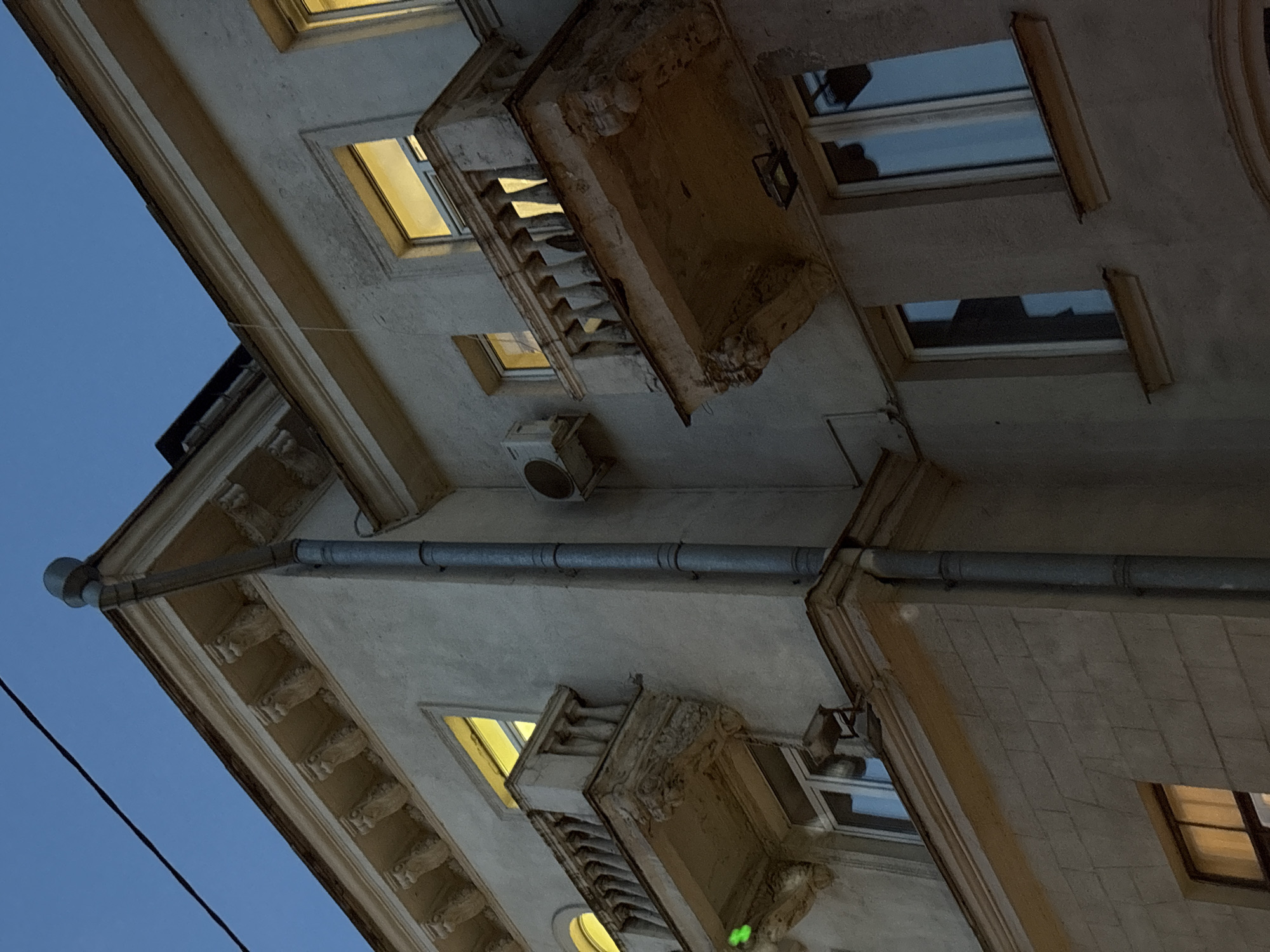 The image is a low-angle shot of a building facade in Minsk, Belarus, taken during the early evening or late dusk. The building features ornate architectural details, including a decorative cornice and balustraded balconies. The exterior is painted in a light color, possibly beige or pale grey. Some windows are illuminated with warm light. A metal gutter runs along the side of the building. The sky is a deep blue. An air conditioning unit is visible mounted on the building exterior. The photo is angled upwards.