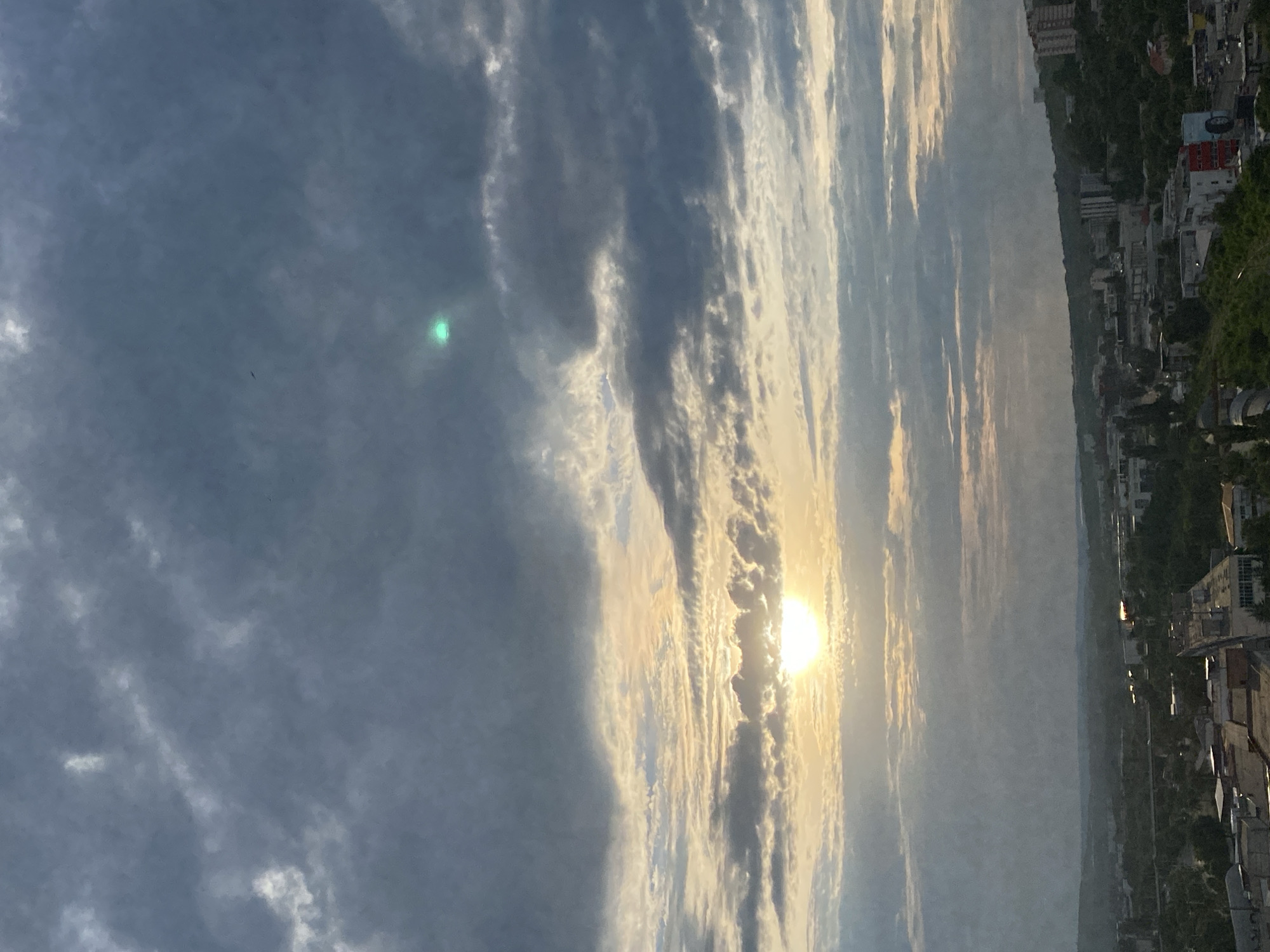 The image shows a wide, vertical shot of a cloudy sky with the sun setting or rising. The sun is a bright, glowing orb partially obscured by textured, wispy clouds that stretch across the frame from left to right. The sky transitions from a darker, overcast gray on the left to brighter, golden hues near the sun, indicating the time of day. Below the sky, a distant cityscape is visible on the right, with buildings and trees lining a hillside. The overall impression is one of a dramatic and serene natural phenomenon occurring over a populated area. There are no people or specific objects in the foreground. The weather appears partly cloudy with the sun breaking through. The location is suggested to be Chișinău, Moldova, which aligns with the distant, somewhat European-looking urban landscape.