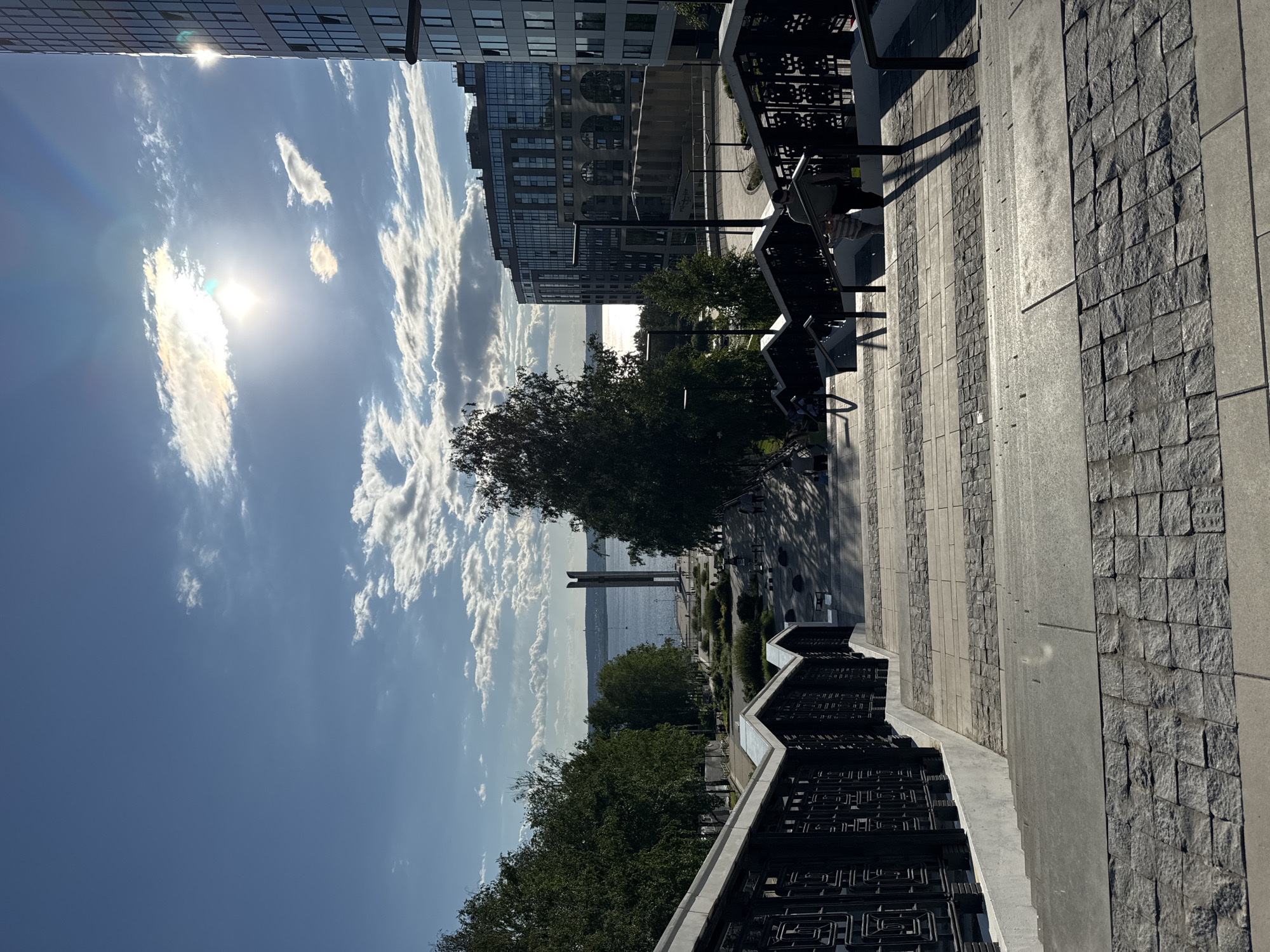 This is a wide-angle, eye-level, outdoor photograph taken on a sunny day, looking down a wide, tiered stone staircase towards a body of water and a city skyline. The staircase is bordered by ornate black metal railings. In the foreground, the steps are constructed of large, light gray concrete slabs interspersed with sections of smaller, dark gray cobblestones.
Further down the staircase, there are several people visible. Two individuals are walking down the steps on the right side, casting long shadows. Other people are scattered throughout the plaza at the bottom of the stairs.
The plaza area features landscaping with trees and bushes, benches, and lampposts. Beyond the plaza is a wide expanse of water, likely a river or lake. On the far side of the water, a tall, slender monument with a distinctive T-shaped top stands prominently.
In the background, a modern city skyline is visible, dominated by several multi-story buildings with glass facades. The building on the far right is particularly tall and sleek. The sky is blue with scattered white clouds, and the sun is visible, creating lens flare and highlighting the scene with bright light. This scene is located in Izhevsk, Russia, specifically at the "Kalashnikov Memorial" or "Victory Park" area along the Izhevsk Pond.