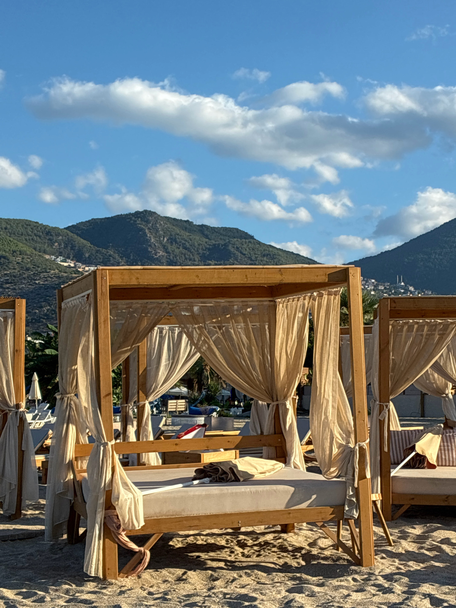 The image shows a beach scene in Alanya, Turkey, under a bright blue sky with scattered clouds. In the foreground, there are several wooden cabanas with draped beige curtains, set up on a sandy beach. These cabanas appear to be lounge beds or resting areas. One cabana in the center is prominently featured, with a beige mattress and a rolled-up blanket or towel on it. The sand in front of the cabanas shows footprints and is textured by the sunlight. Behind the cabanas, there are more lounge chairs, umbrellas, and some tropical vegetation. In the background, lush green mountains rise up, with buildings visible on the slopes and a prominent white-domed structure, likely a mosque, atop one of the hills. The overall impression is one of a peaceful and inviting coastal resort setting.