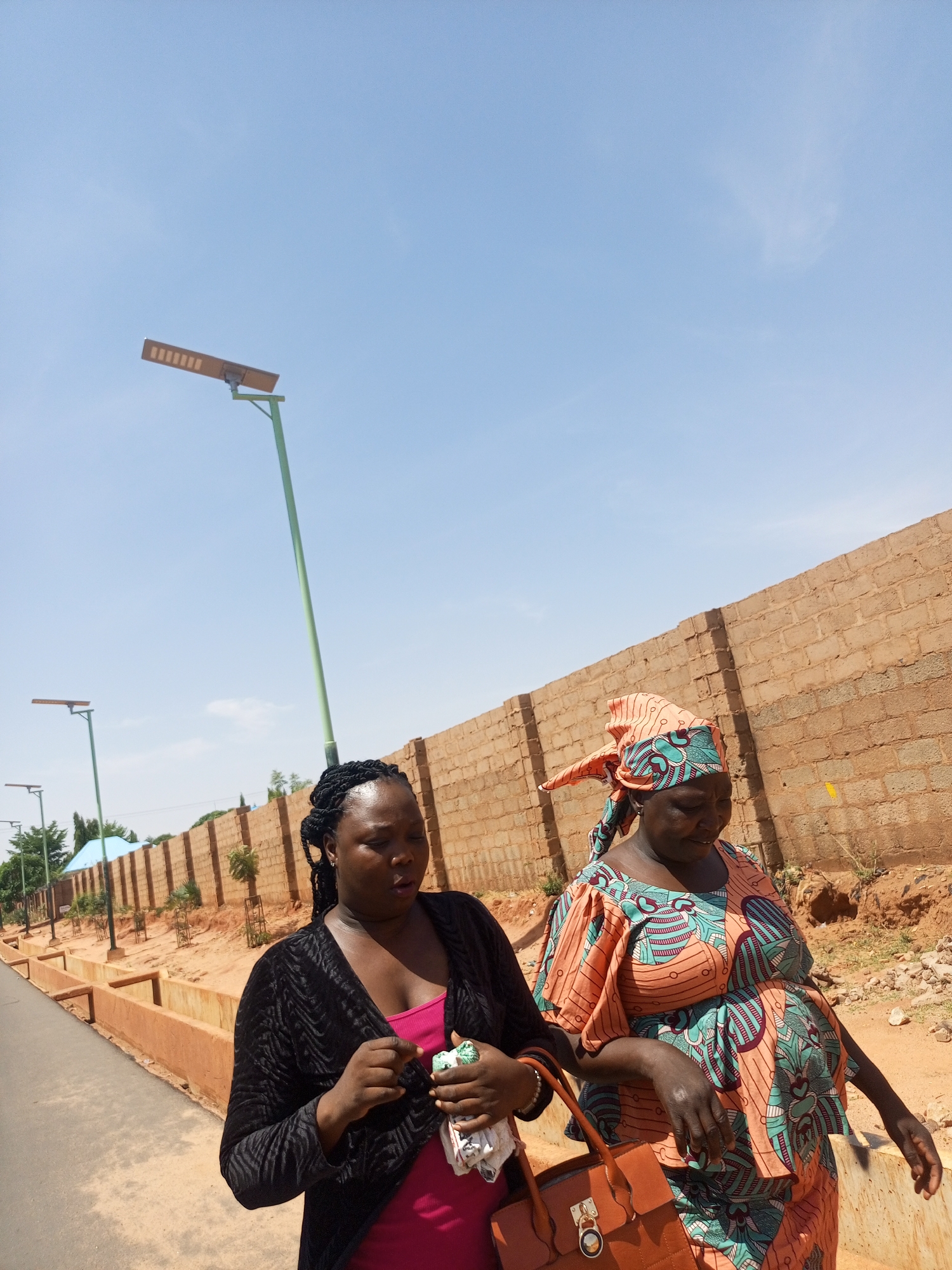 Two adult women walk along a paved road beside a brick wall, with intermittent street lights on green poles. The woman on the left wears a pink top and a black jacket, and holds a white cloth or tissue. The woman on the right wears a peach and teal patterned dress and a matching head wrap, and carries a brown handbag. The sky is clear and blue, and the location appears to be a city street in Unknown, Unknown.