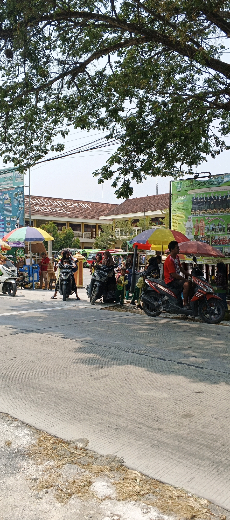 The image depicts a street scene in Karangwungu, Indonesia, on a sunny day. In the foreground, a grey concrete road with textured markings is visible, bordered by dry grass and debris on the left. The middle ground features several motorbikes, some with riders. On the right, a man in a red shirt is on an orange motorbike. To his left, under a red and yellow umbrella, a woman in a headscarf is sitting on a black motorbike. Further left, two children in green uniforms are standing near a stall. Behind them, a black motorbike carries two people, and to its left, another motorbike has a rider. In the background, a building with a tiled brown roof, identified by the text "SMHD HASYIM MODEL" on its facade, is prominent. Two banners hang in front of the building. The overall impression is that of a bustling area, possibly near a school or marketplace, with daily activities taking place on the street.