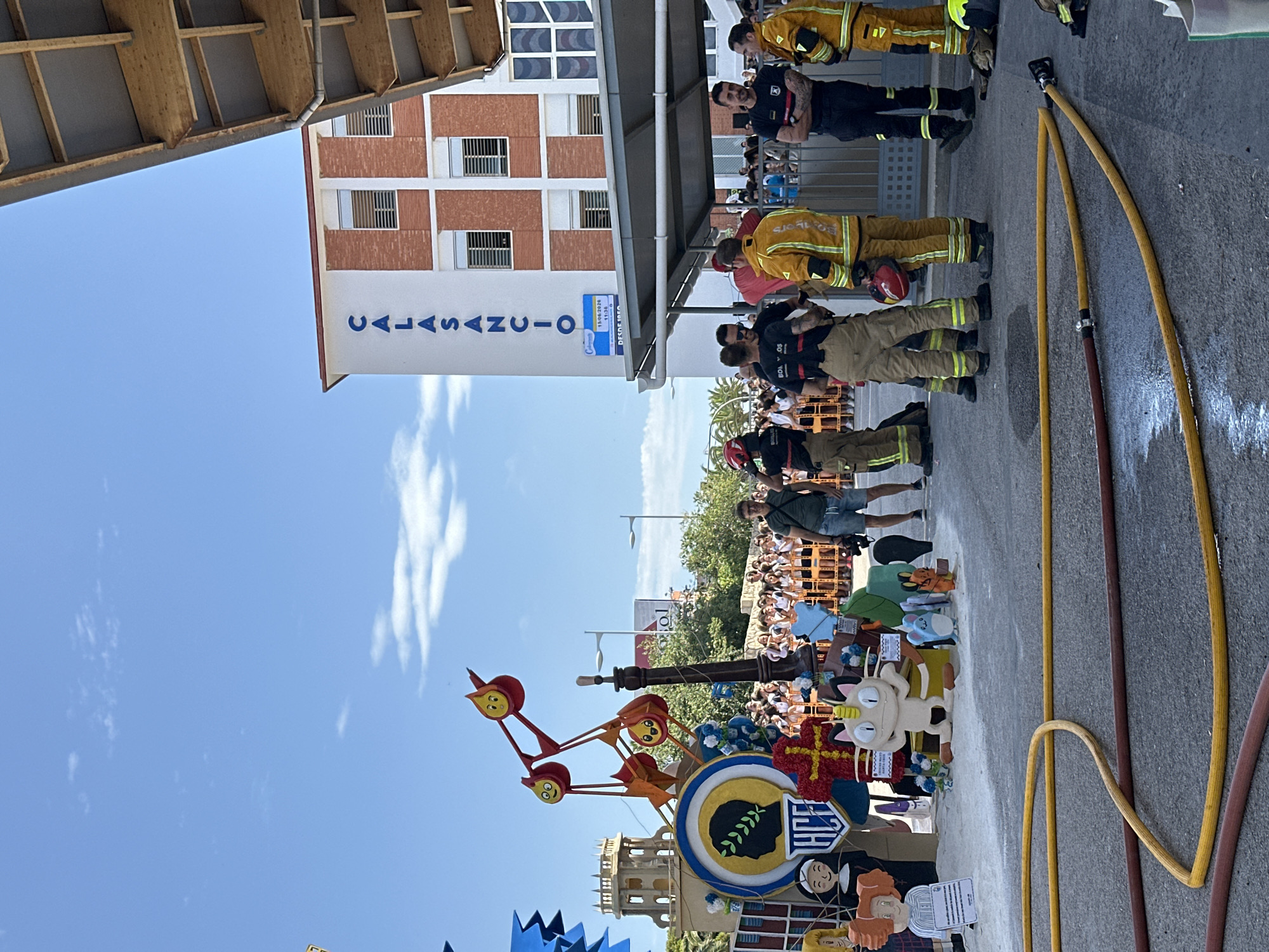A medium shot captures an outdoor scene with a building labeled "CALASANCIO" in blue letters against a white and red brick facade. The building has several windows with blinds. In the foreground, a group of firefighters in yellow and tan uniforms stand and converse. Fire hoses, one yellow and one red, lie on the asphalt in front of them. Behind the firefighters, a colorful display of large, animated figures and structures is visible, including cartoonish characters and a depiction of a Roman-style bust. A crowd of onlookers stands behind barriers in the middle ground. The sky is blue with a few scattered clouds. The overall impression is that of a public event or festival.