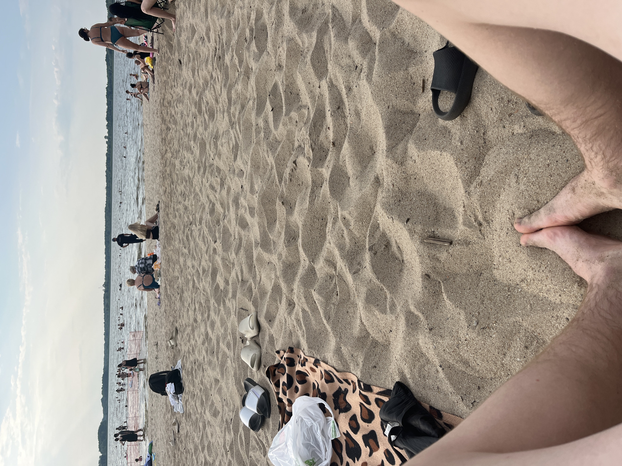 The image depicts a sunny day at a crowded beach, likely in Загародскі сельскі Савет, Belarus. The scene is dominated by sand, with the water of a lake or river in the background, where many people are swimming and enjoying the water. On the sand, several individuals are scattered, some sitting, some lying down, and others walking. There are various objects present, including towels, beach bags, and footwear, suggesting a day of leisure and recreation. The weather appears to be warm and pleasant, with clear skies and the sun casting shadows on the sand, indicating it is daytime. The overall atmosphere is one of a lively and active beach environment on a summer day.