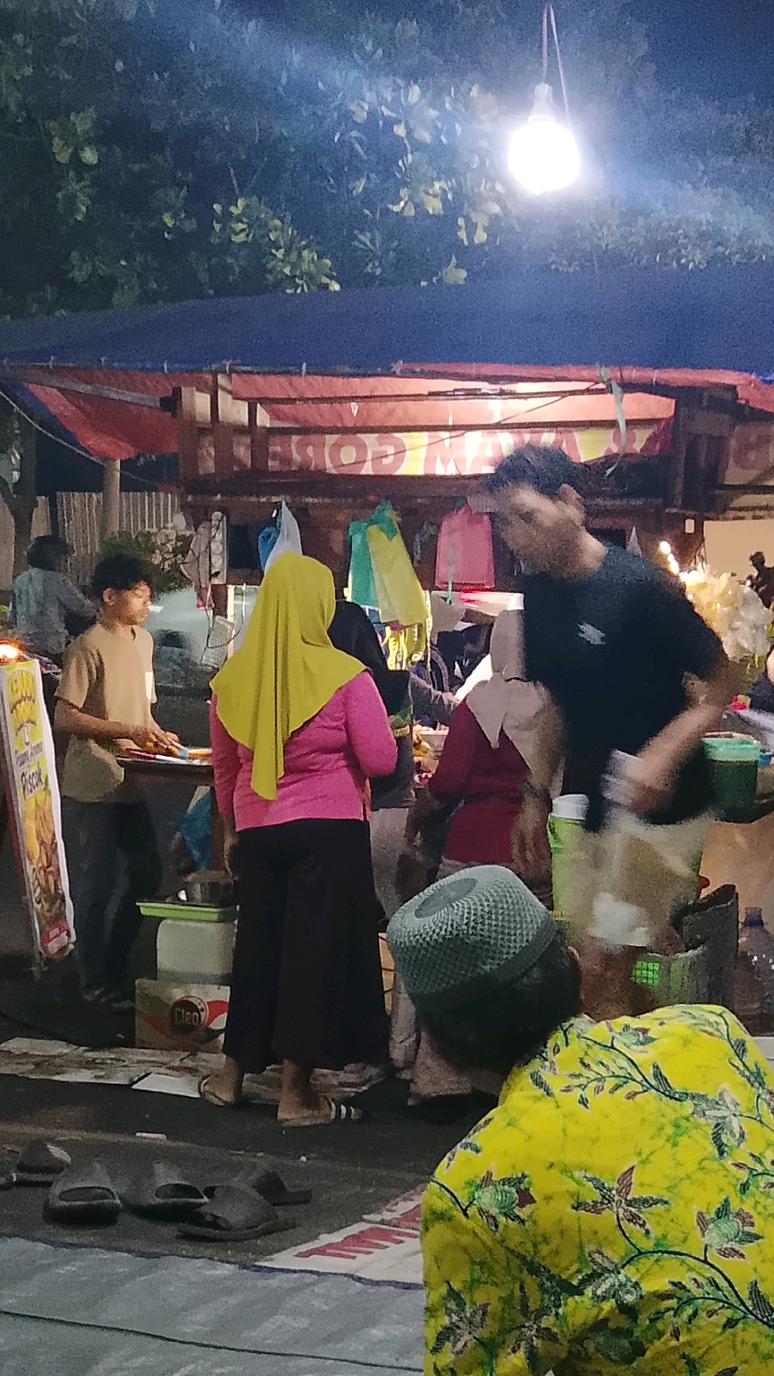 In Randuagung, Indonesia, at night, a street food stall is illuminated by a bright overhead light. Several people are visible. A young man in a tan t-shirt stands behind a table, appearing to be preparing or serving food. A woman wearing a bright yellow hijab and a pink shirt stands facing away from the camera. To her right, a person in a dark shirt with a blurry motion is interacting with someone to their left, who is partially visible and wearing a light-colored headscarf and a red garment. Further back, a person in a light blue shirt is seated. Scattered on the ground are several pairs of sandals and mats. The stall has a blue tarp overhead and displays various items of clothing, some brightly colored. Signs and containers are also present, with one sign reading "Piscok." The overall scene suggests a bustling night market environment.