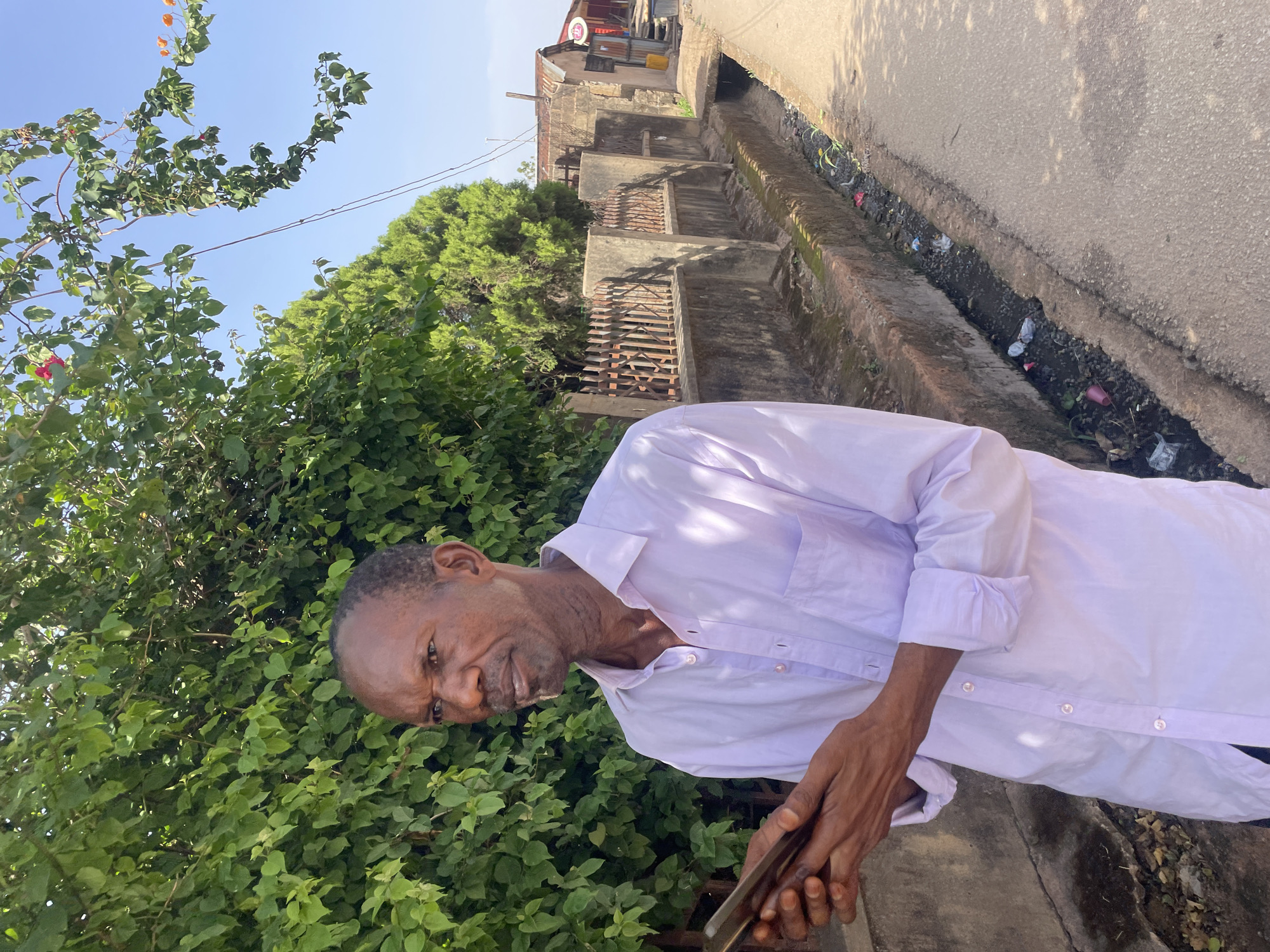 A man wearing a long-sleeved white shirt stands on a paved road next to a drainage ditch in Jos, Nigeria. He is facing the camera and holding a mobile phone. Behind him is dense green foliage. To the right, the drainage ditch runs alongside the road, filled with debris. Beyond the ditch are houses and buildings, suggesting a residential area. The sky is partially visible, indicating daytime.