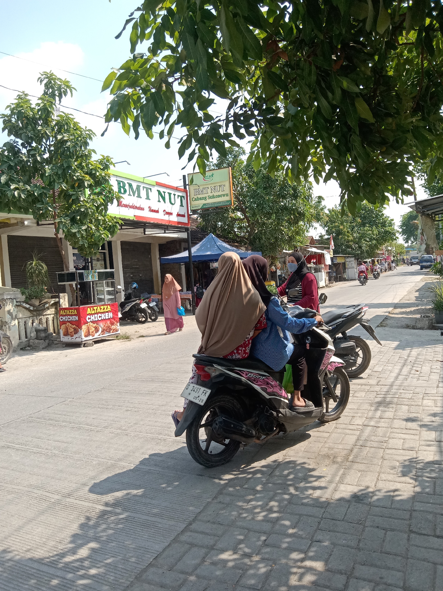 In the city of Klepek, Indonesia, on a sunny day, a group of people are seen on a street with a sidewalk. A woman in a pink hijab and a blue bag walks past two motorcycles parked on the side of the road. Two individuals are riding on one of the motorcycles, their faces covered by hijabs. Another woman in a red shirt and a black mask is seen on the back of a separate motorcycle, also with a hijab.

In the background, a blue tent is set up, and several other motorcycles and a van are visible on the street. Buildings line the street, one of which has a sign for "BMT NUT" and another smaller sign with "ALTAYZA CHICKEN" and images of fried chicken. Trees with green leaves provide shade over the scene. The ground is paved with concrete and cobblestones, with shadows cast from the trees.