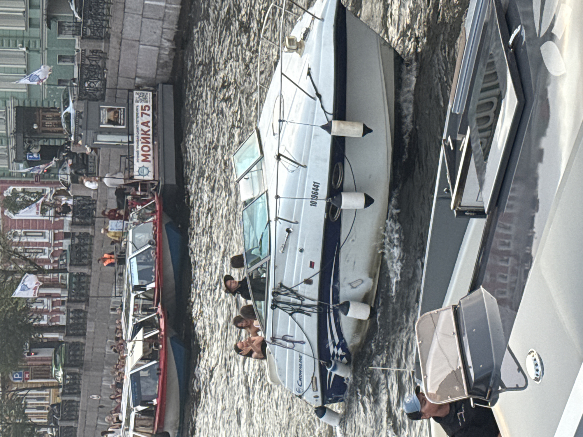 This image shows a bright, sunny day on a river in Saint Petersburg, Russia. The scene is dominated by several boats on the water. In the foreground, a sleek, white motorboat with blue trim is visible, its cabin windows reflecting the surroundings. The boat has a registration number "10B6441" and the brand "Crownline" is visible. Two white fenders are attached to its side. Another larger boat with a grey and white exterior is partially visible to the right, also on the water. Further down the river, a red and blue tour boat is packed with people, suggesting a tourist activity or river cruise. The boat has multiple windows and appears to be a popular mode of transport. The background reveals the city's architecture, with buildings lining the riverbank. There are people visible on the boats, some appearing to be passengers enjoying the ride and the scenery. One person on the motorboat is wearing a cap. The overall atmosphere seems lively and pleasant, consistent with a sunny day in a tourist city. A sign on the riverbank reads "РЕЧНОЙ ТУРИЗМ. САНКТ-ПЕТЕРБУРГ. МОЙКА 75", indicating river tourism in Saint Petersburg at Moika 75. The water appears calm with ripples.
