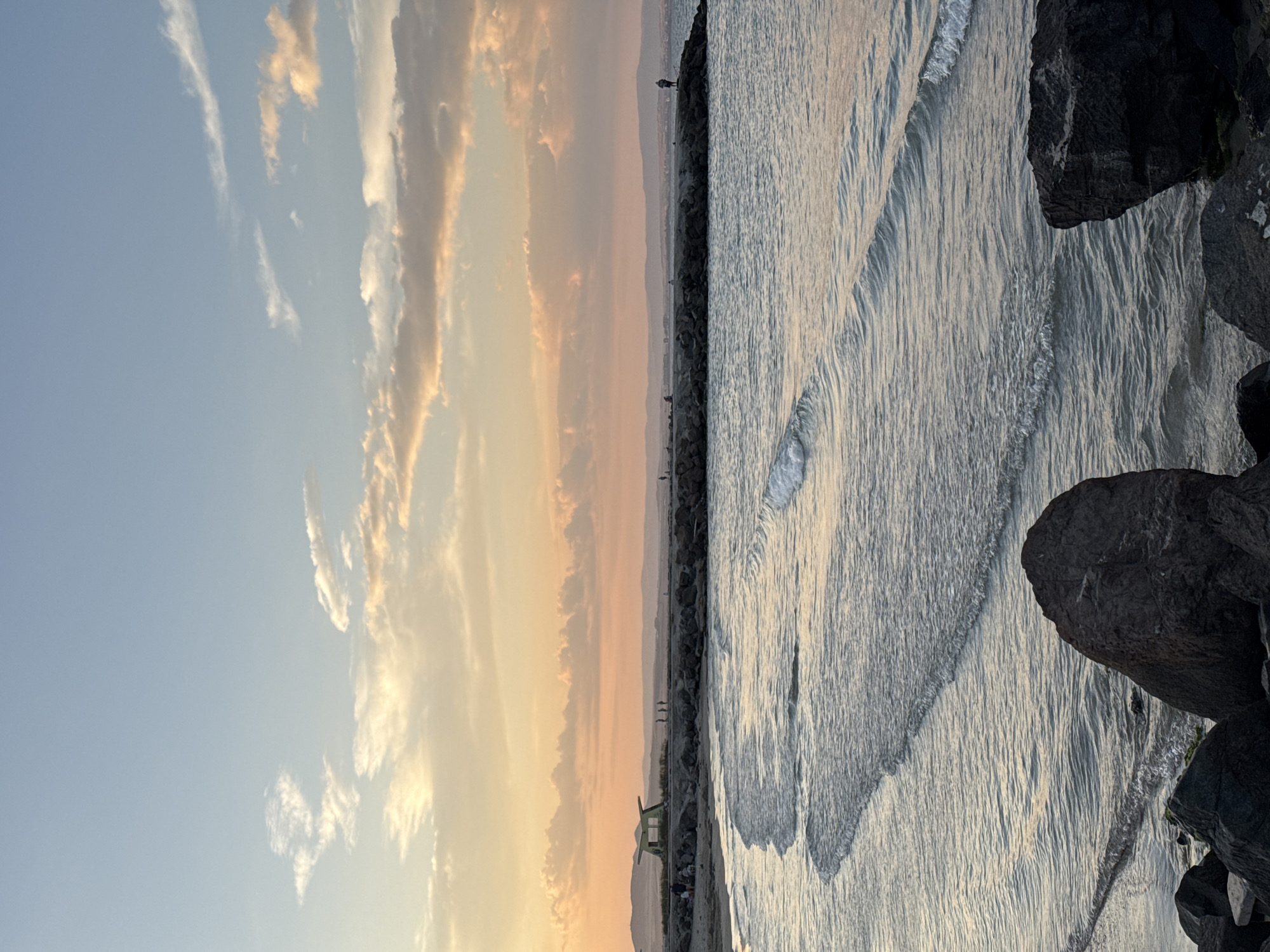 The image depicts a coastal scene at sunset in Pomorie, Bulgaria. The sky is a gradient of blue at the top, transitioning to warm hues of orange and pink from the setting sun, with scattered clouds reflecting the light. In the foreground, large dark rocks are situated on a beach or shoreline. The sea is calm with gentle waves lapping at the shore. A stone pier extends into the water, with several silhouetted figures walking on it. One figure stands at the end of the pier, appearing to fish or observe the sea. Further down the coastline, a small, light-colored structure with a green roof is visible near the beach, where a few more figures are gathered. The overall impression is one of tranquility and natural beauty.
