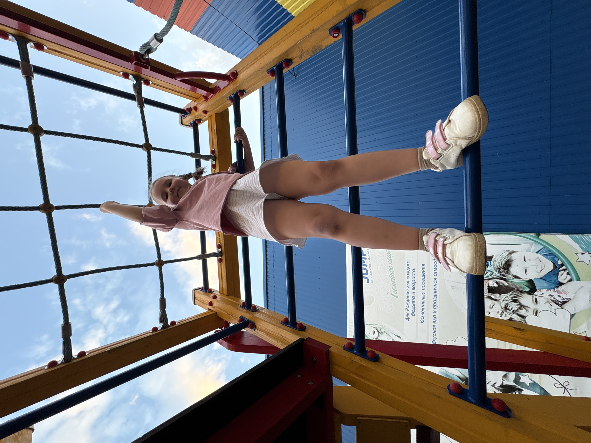 A young girl is seen playing on playground equipment, specifically a combination of rope netting and monkey bars. She is positioned horizontally, appearing to be in motion or maintaining balance on the apparatus. The location appears to be in Chaikovsky, Russia, as indicated by the Russian text and logo for the "Jump" trampoline park visible on a wall or sign near the playground structure.