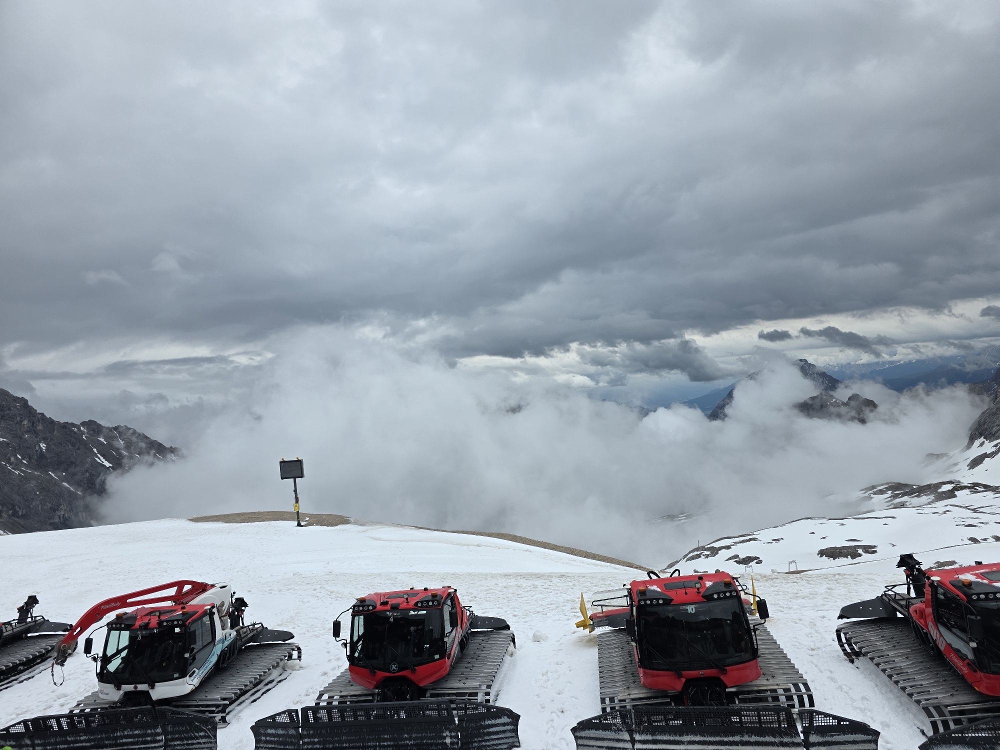 The image captures a high-altitude, snow-covered landscape on the Zugspitze glacier, near Garmisch-Partenkirchen, Germany, under a heavily overcast sky. A dense layer of grey stratus and cumulus clouds dominates the upper half of the frame, with a "sea of clouds" partially obscuring the lower mountain ranges and distant valleys. Hints of darker, jagged mountain peaks with patches of snow are visible through and above the cloud layer, particularly to the far left and in the background.
In the foreground, five stationary snow groomers (PistenBully models), predominantly red and black, are parked on a wide expanse of compacted snow. From left to right: a partially visible groomer with a red articulated crane-like attachment, a full red and black groomer marked with "4", a full red and black groomer, a groomer with a large yellow rear attachment (likely a tiller or blade) marked with "10", and a partially visible groomer on the far right. All groomers feature wide metal track systems. Black protective netting or fencing is visible in the extreme bottom foreground. In the mid-ground, a black square information board mounted on a yellow pole stands on a patch of snow-free, rocky ground. No individuals are present in the scene.